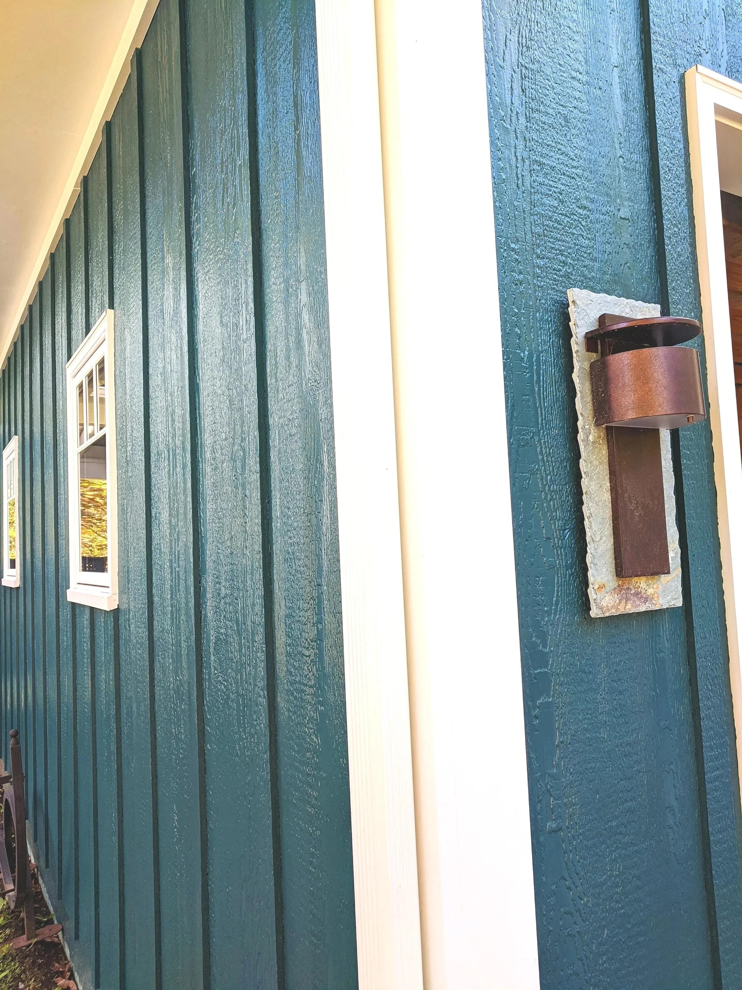 Close-up of the exterior of a house painted in teal with vertical wooden siding, white framing around a window, and a rustic wall-mounted light fixture.