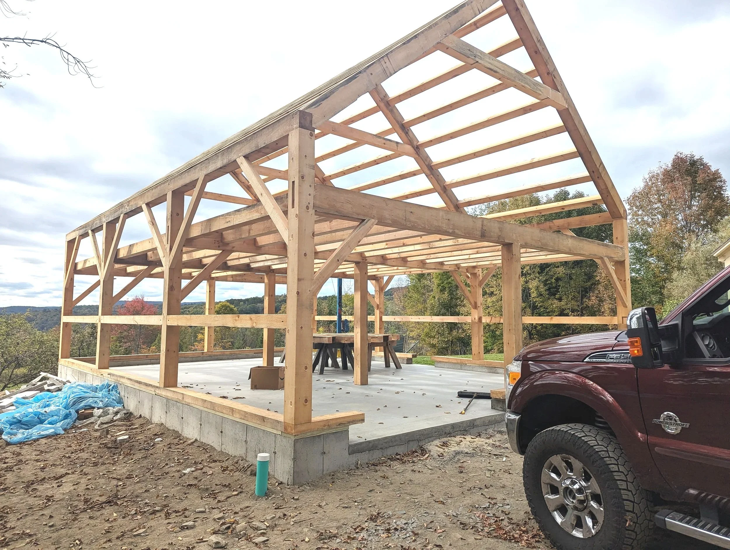 Construction site with a wooden structure frame on a concrete foundation, partly covered roof, outdoors in a rural area with trees in the background, and a maroon truck parked nearby.