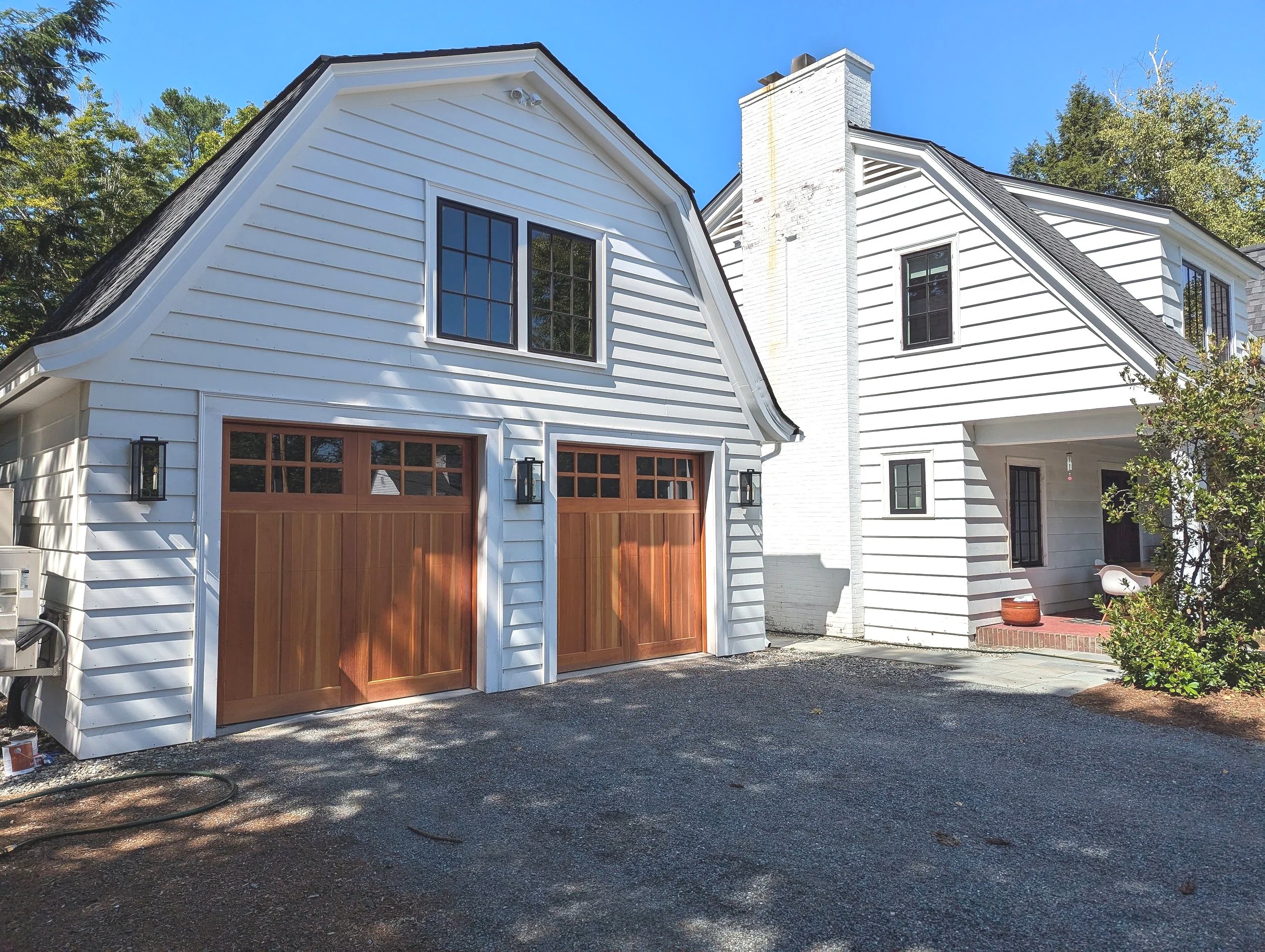 A white house with a garage that has wooden doors and black lantern-style lights on either side. The house has a steep black roof, multiple windows, and a white brick chimney. There is a driveway and some greenery around the house.
