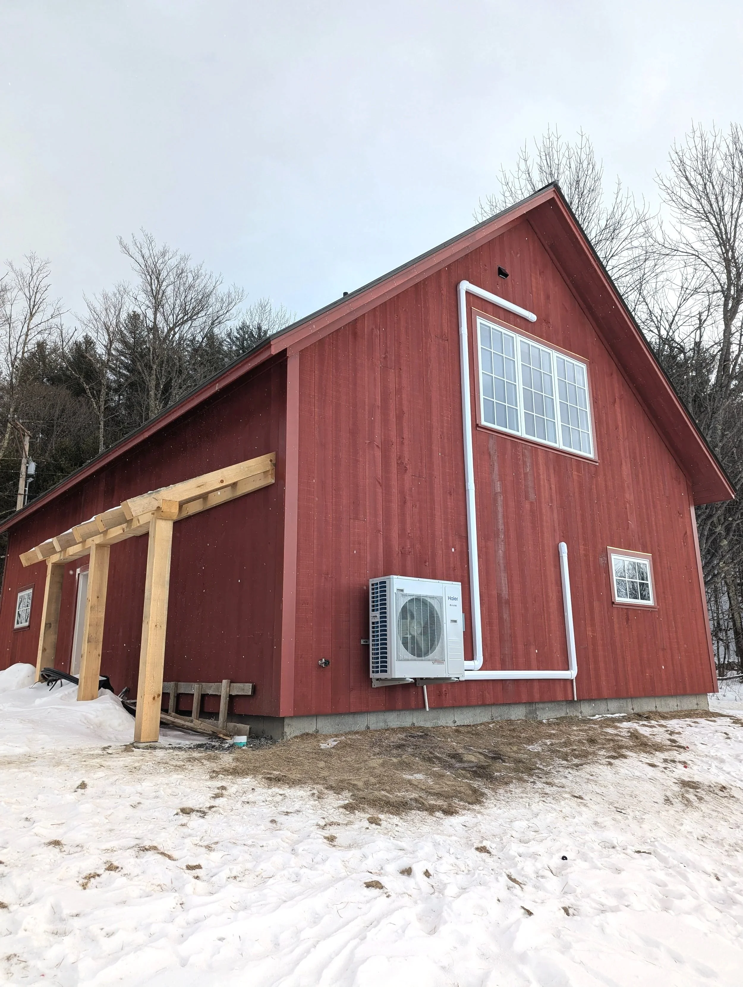 Red barn with solar window panels, HVAC unit, white drain pipes, and a wooden structure at the side, snow on the ground, trees in the background, overcast sky.