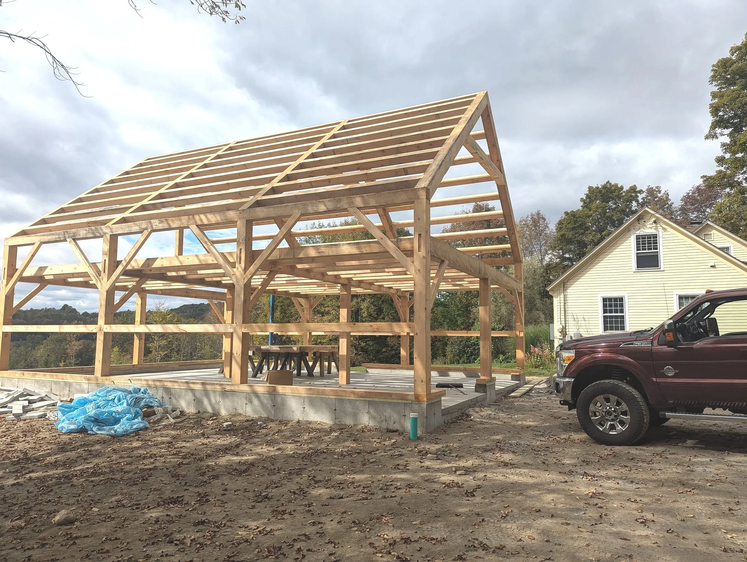 Construction site with a wooden framework for a house or barn, a pickup truck parked nearby, and a residential house in the background on a cloudy day.