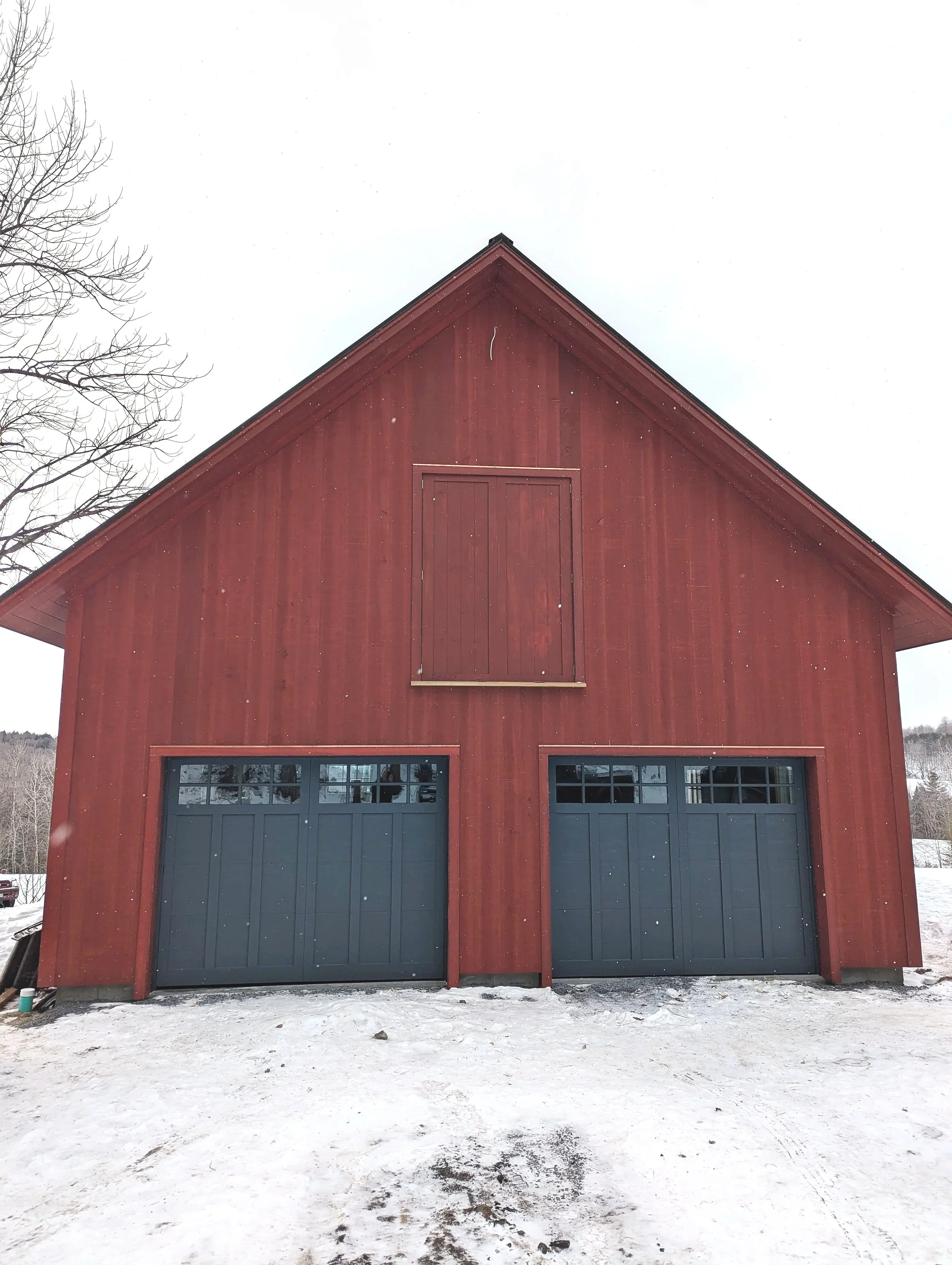 A red barn with closed black garage doors, a vertical barn door above, leafless trees, and snow on the ground.