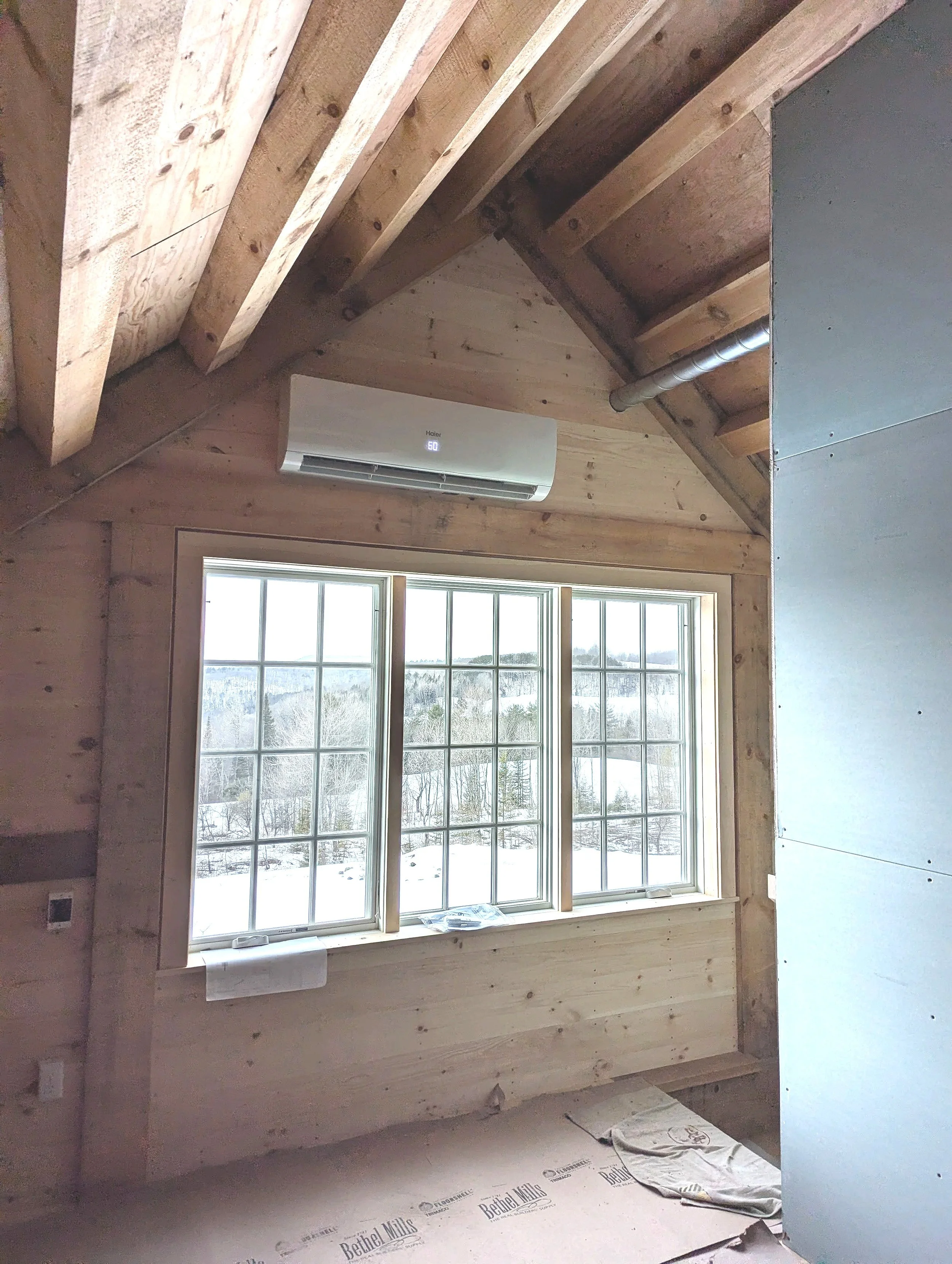Interior of a room under construction with a large window overlooking a snowy landscape, an air conditioning unit on the wall, and exposed wooden ceiling beams.