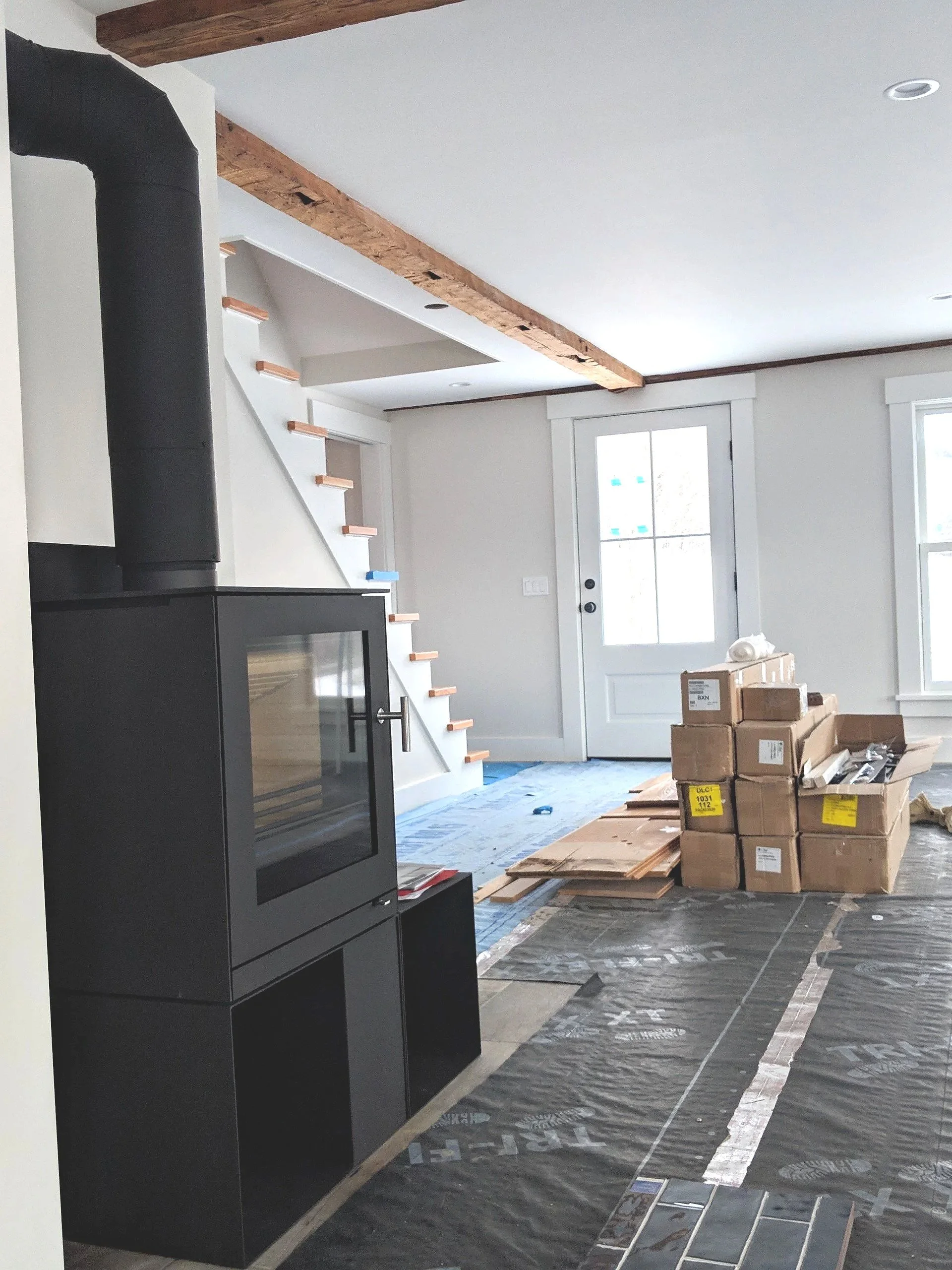 Living room under renovation with flooring installation in progress, boxes and tiles scattered, and a wood beam on the ceiling.