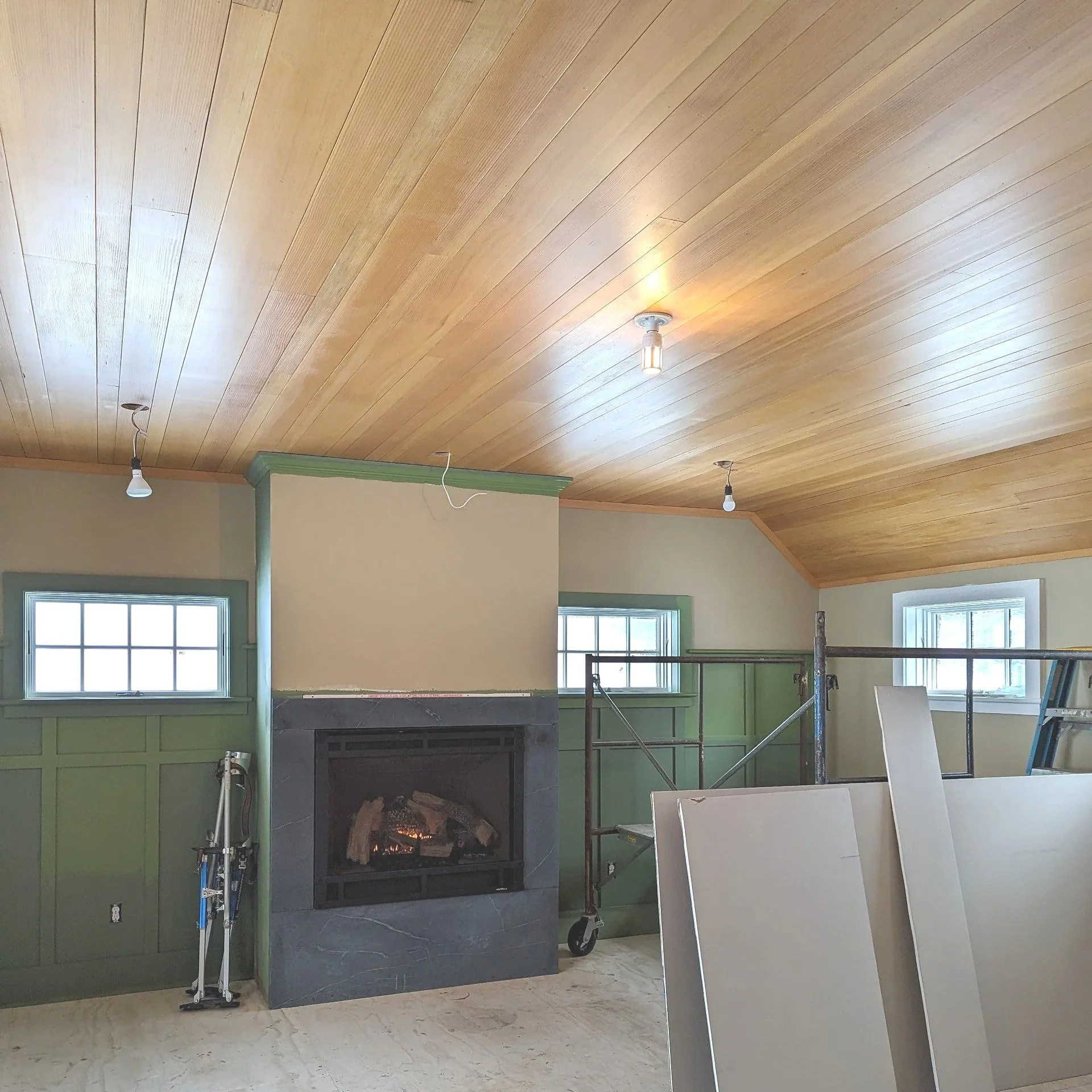 Interior of a room with a wooden ceiling, a fireplace with a black frame, two small windows, a stepladder, and some construction materials.
