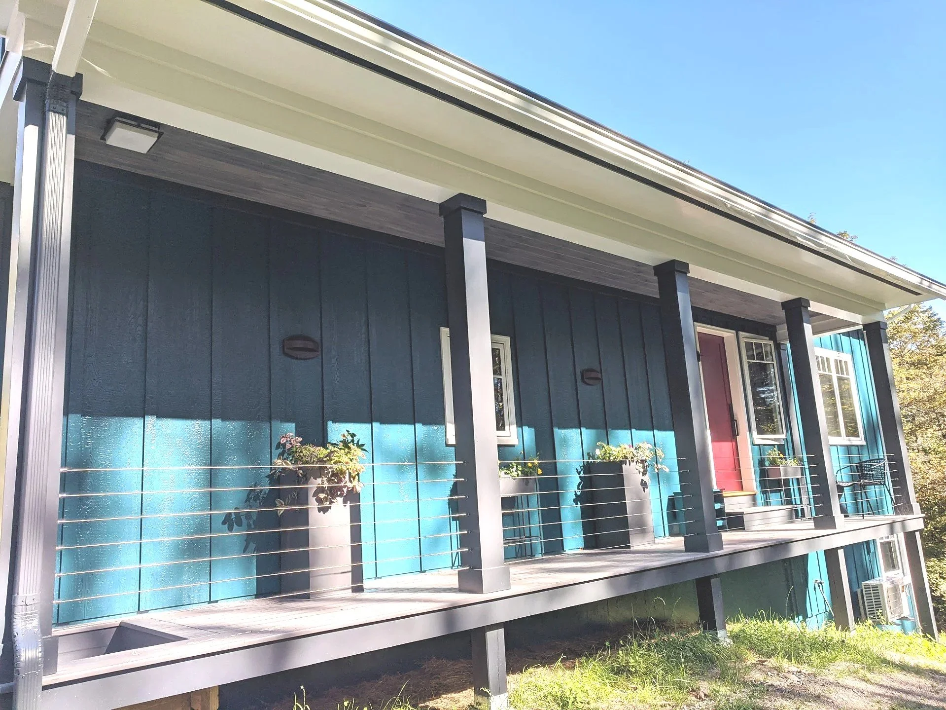 Front porch of a house painted in dark teal with black support beams, two planters with flowers, a red front door, and a white window, with a lawn in front and blue sky above.