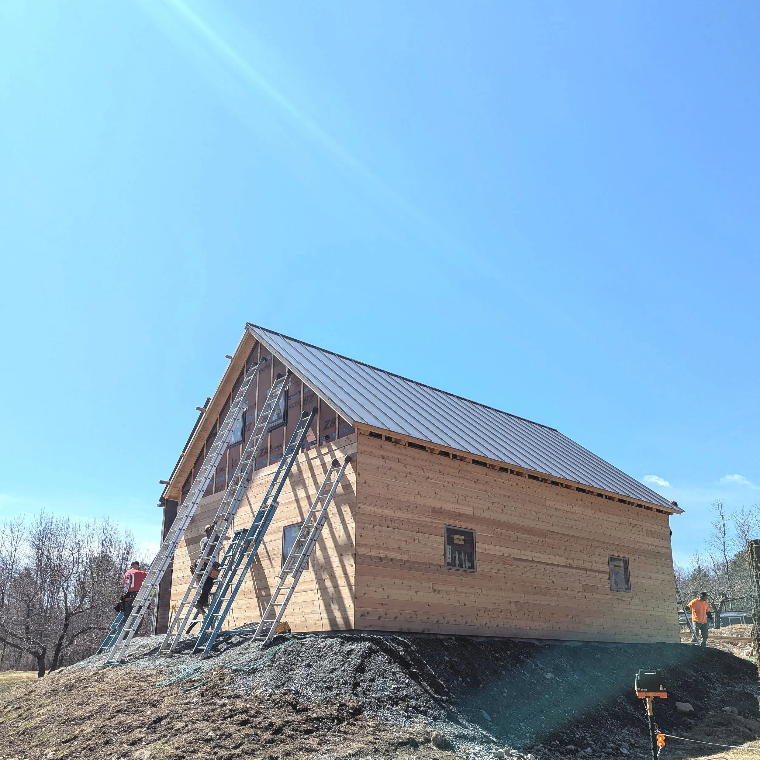 Construction of a wooden house with a metal roof, ladders leaning against it, workers on site, and a clear blue sky.