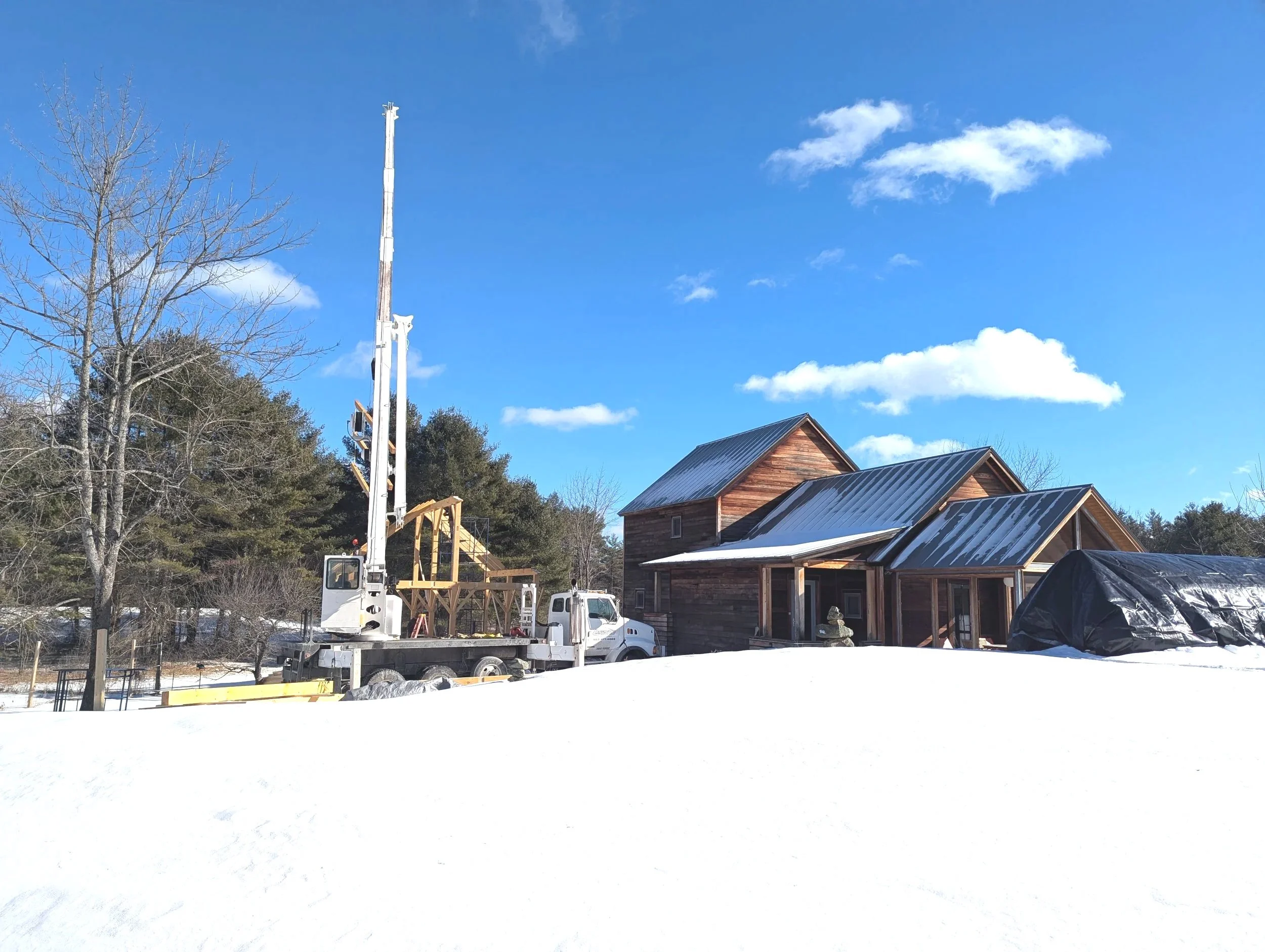 Construction site with a wooden house, snow on the ground, a crane truck, and a blue sky with clouds.
