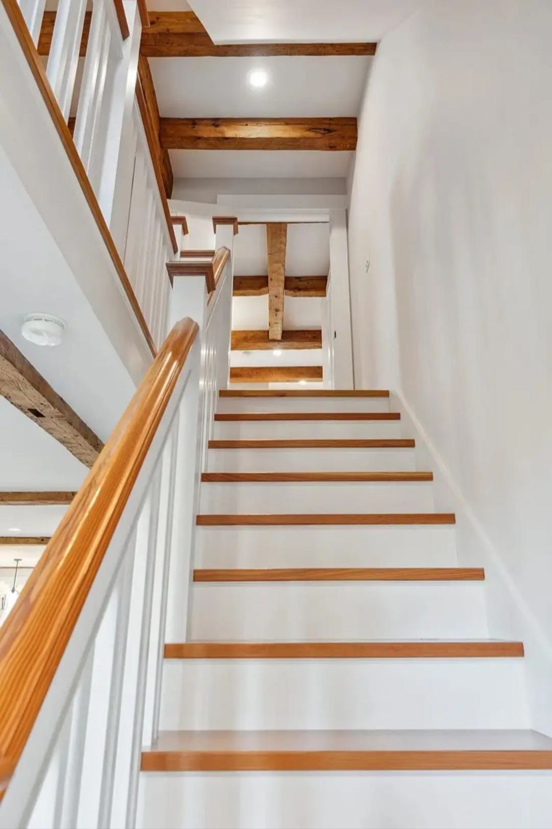 View of a staircase with wooden steps and white risers, leading upward to a second floor with exposed wooden beams on the ceiling.
