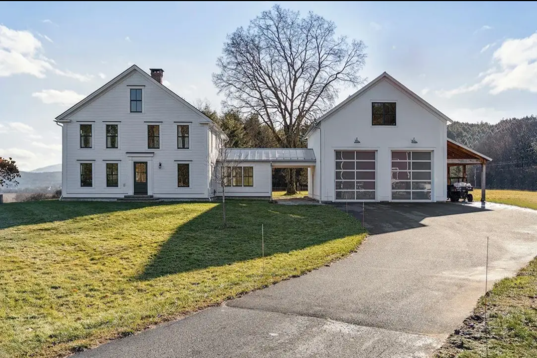 A large white house with multiple windows on the left, a smaller white garage with glass doors on the right, connected by a glass-enclosed walkway. There is a driveway leading to the garage and an open porch area on the right side of the garage with 