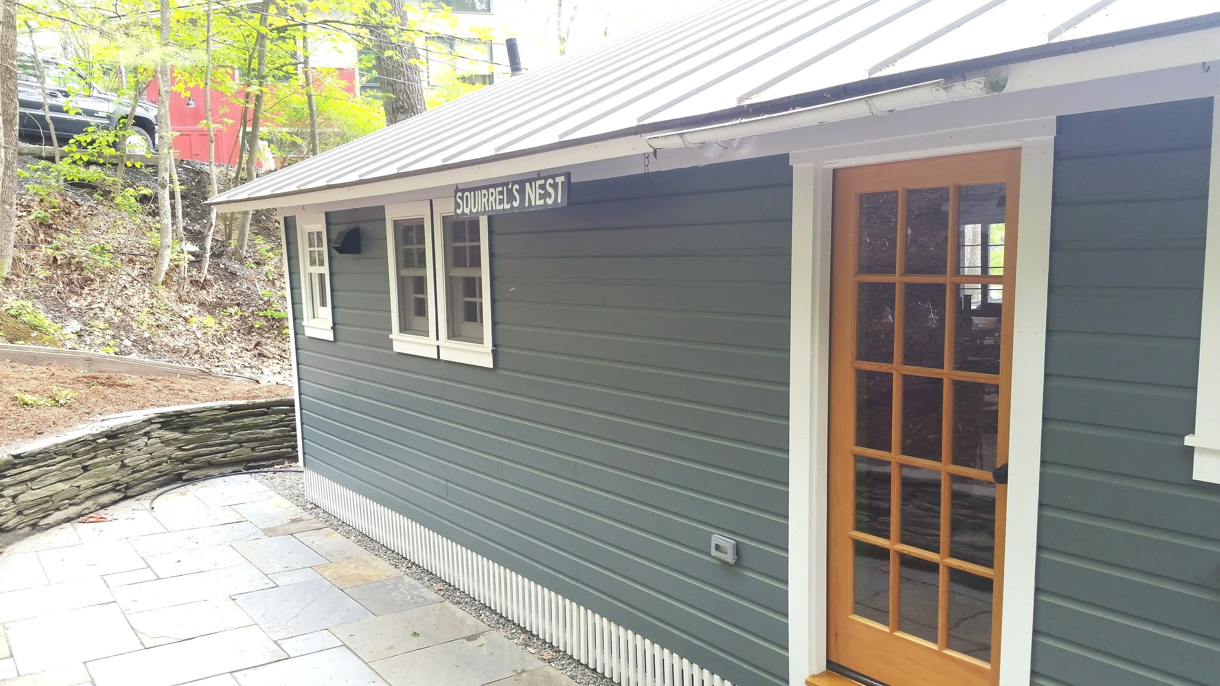 Exterior of a small gray building with white trim, labeled "Squirrel's Nest," featuring three small windows and a wooden door with glass panes, situated next to a stone pathway and surrounded by trees.
