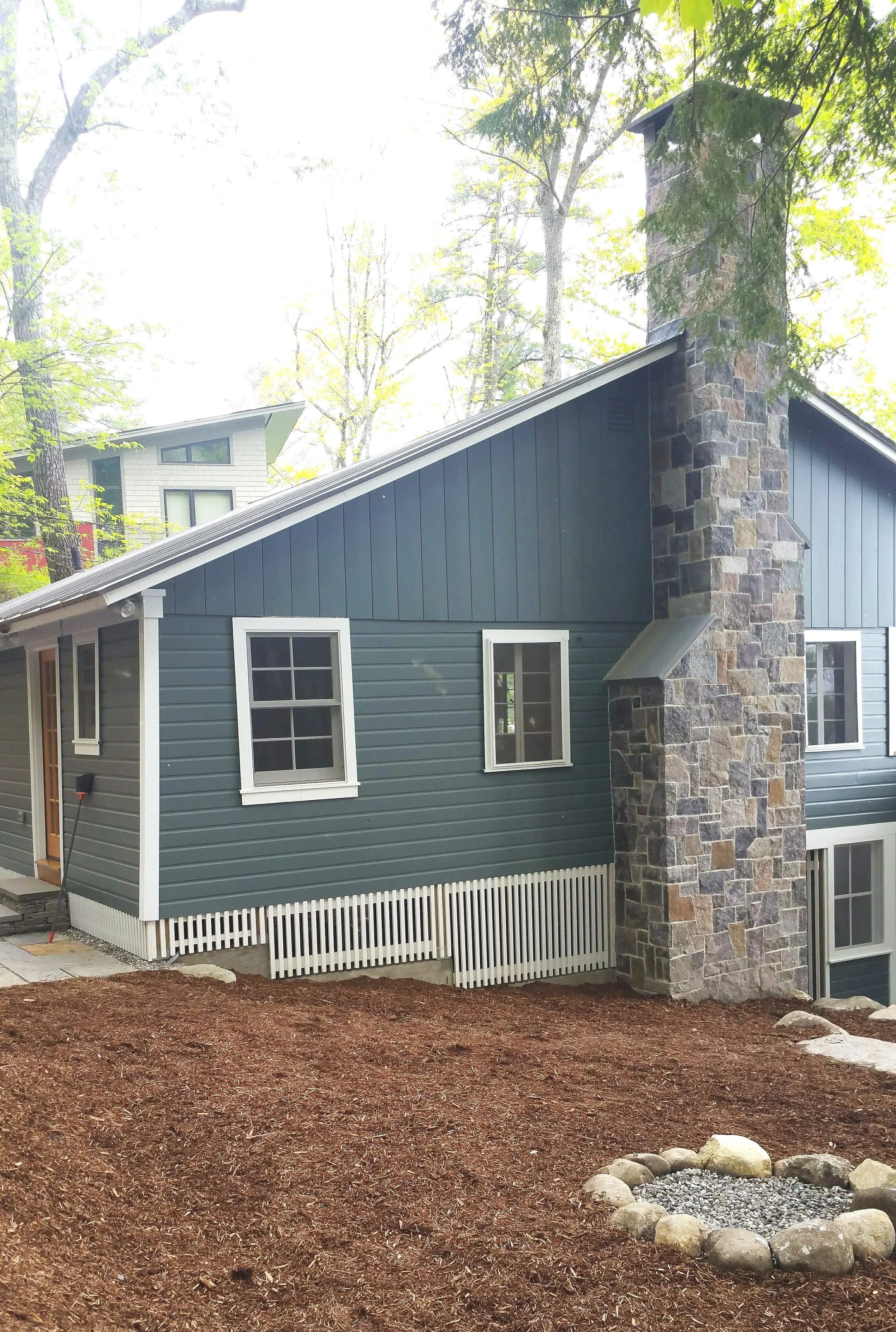 A house with blue siding, white framed windows, and a stone chimney in a wooded area.