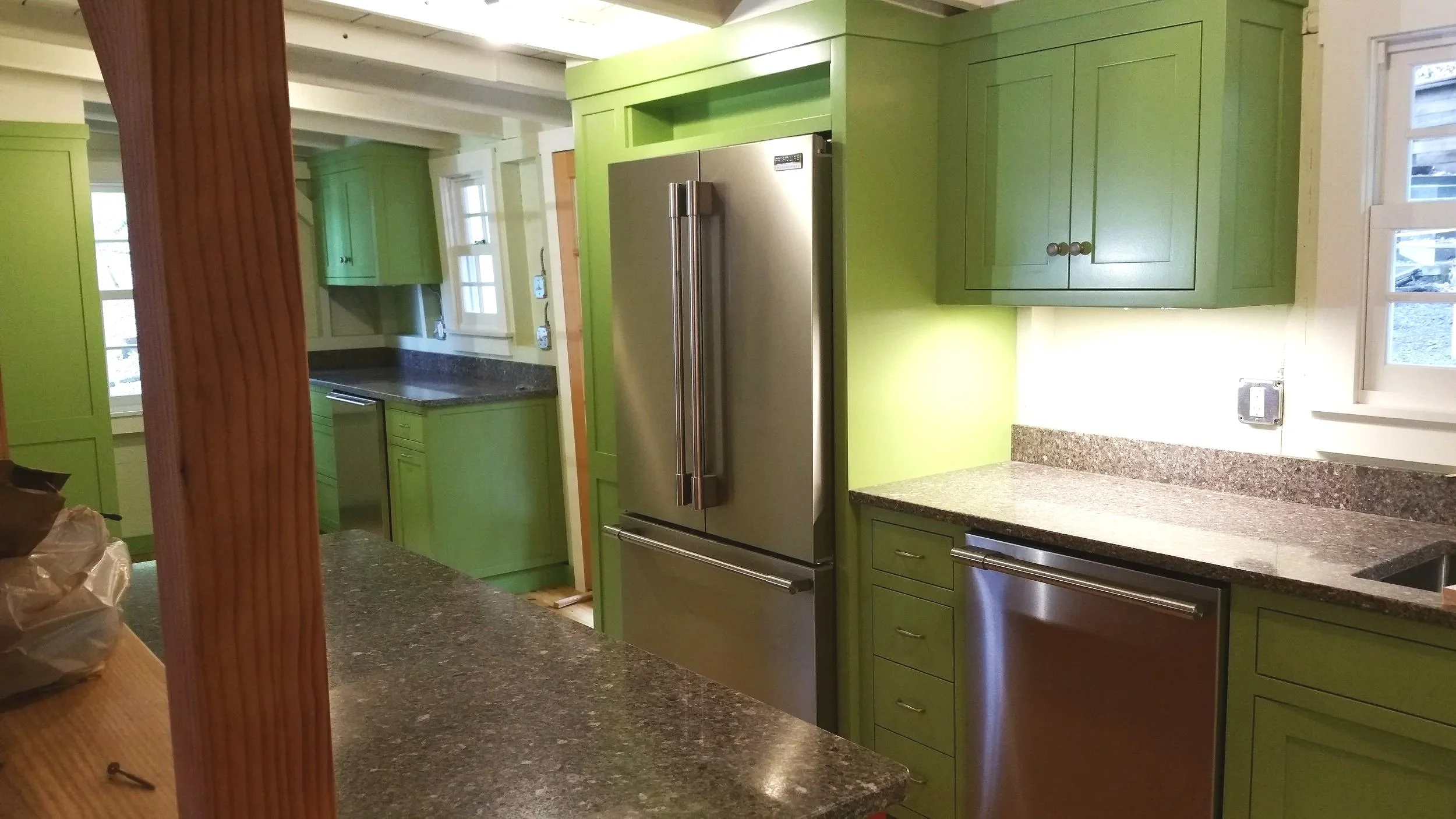 A kitchen with bright green cabinets, granite countertops, a stainless steel refrigerator, a dishwasher, and windows letting in natural light.