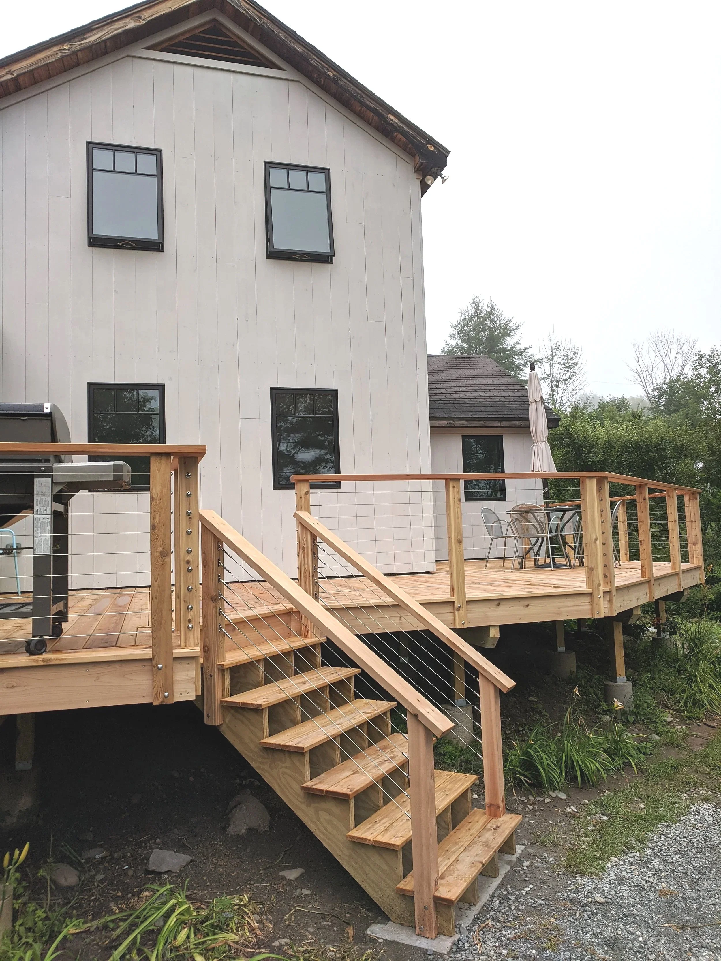 Newly built wooden deck with stairs and railing attached to the back of a white house with four windows, an outdoor table with chairs, and a barbecue grill