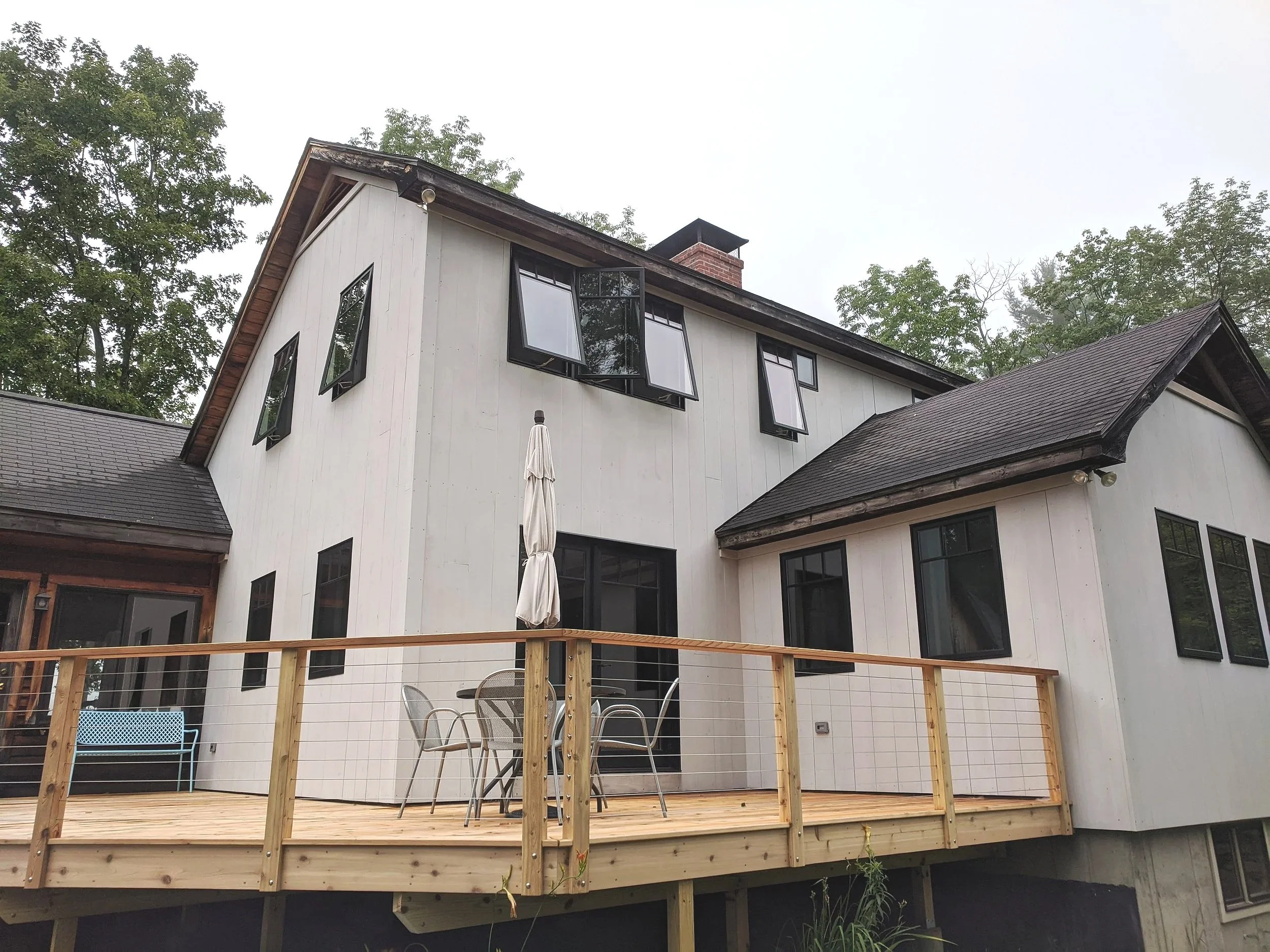 White multi-story house with black window frames and a wooden deck with outdoor furniture, surrounded by trees.