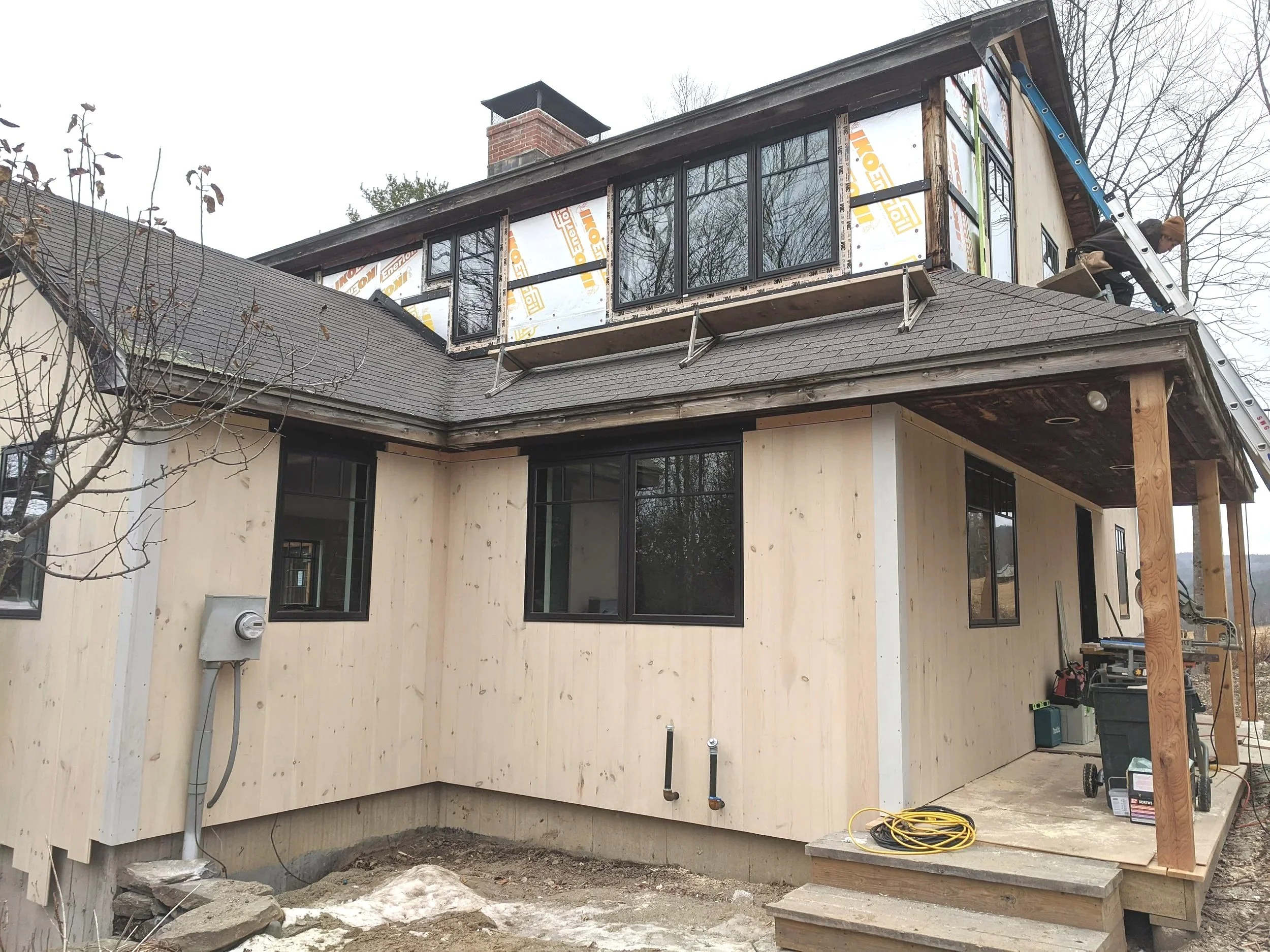 Construction workers building a room extension on a house, with new wooden siding and a window frame, using a ladder to access the upper level.