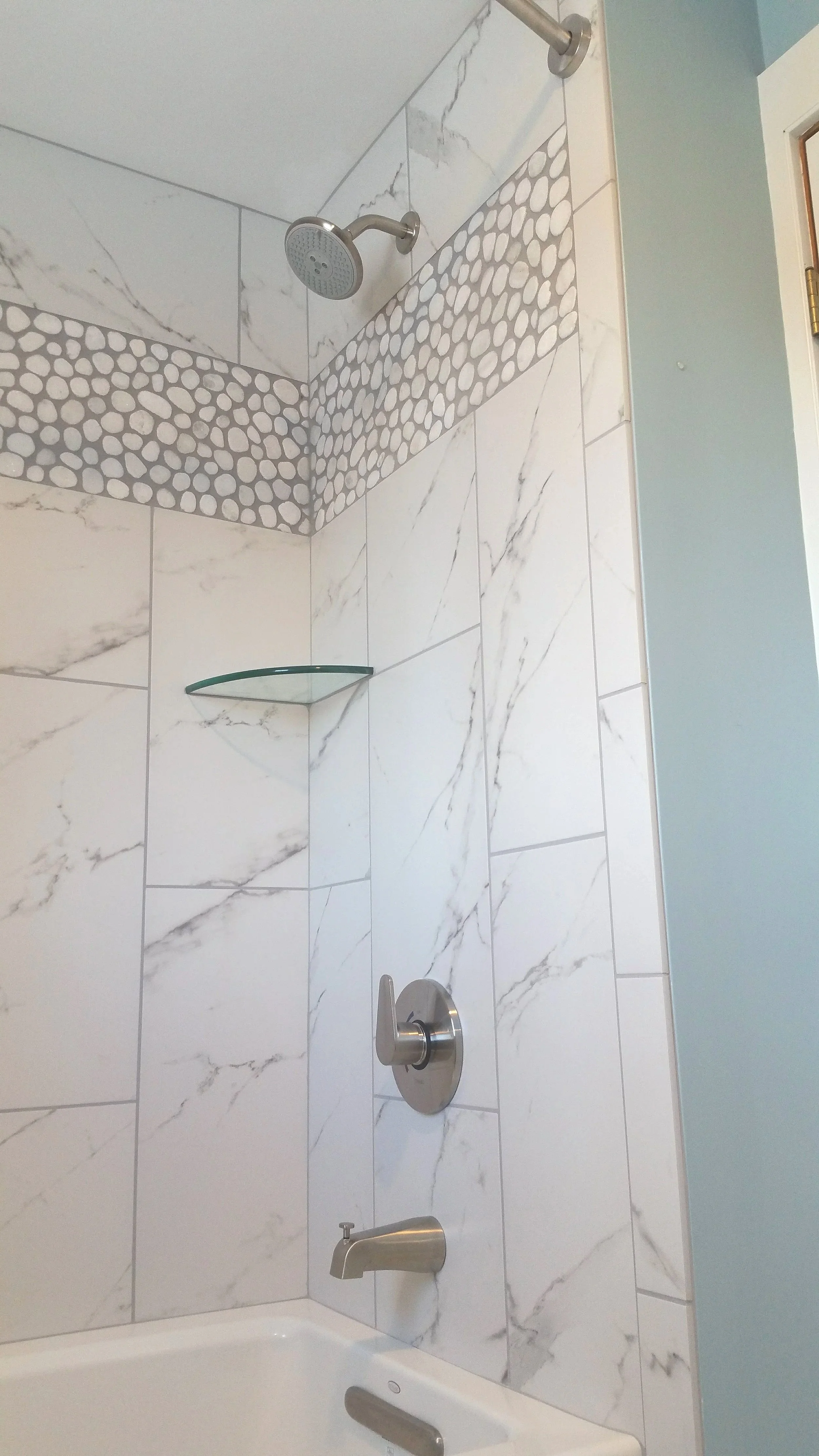 Close-up of a bathroom shower with white marble tiles, a glass corner shelf, a silver showerhead, a silver faucet, and a silver knob.