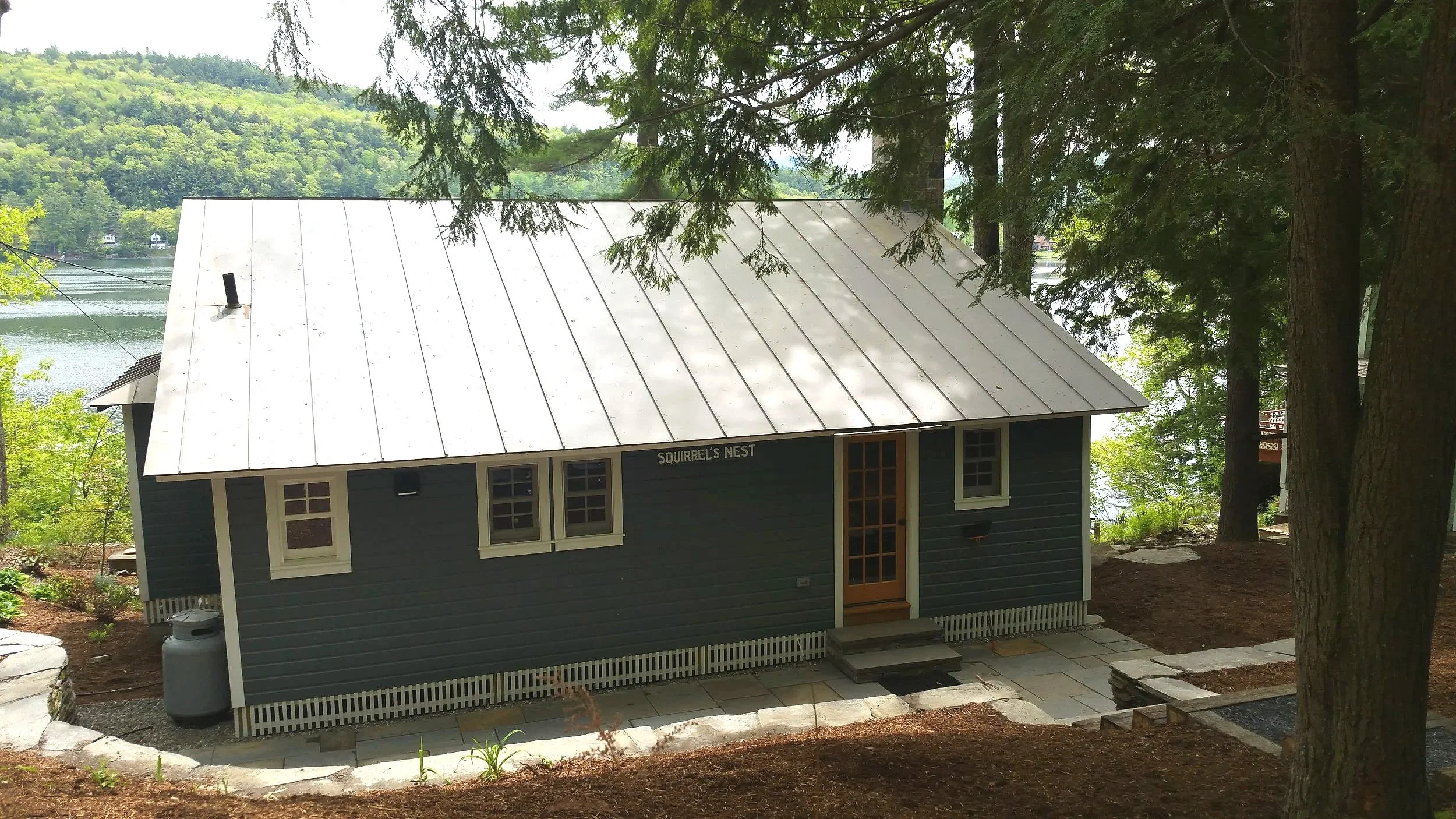 A small blue building named 'Squirrel's Nest' with a white metal roof, situated among trees near a body of water.