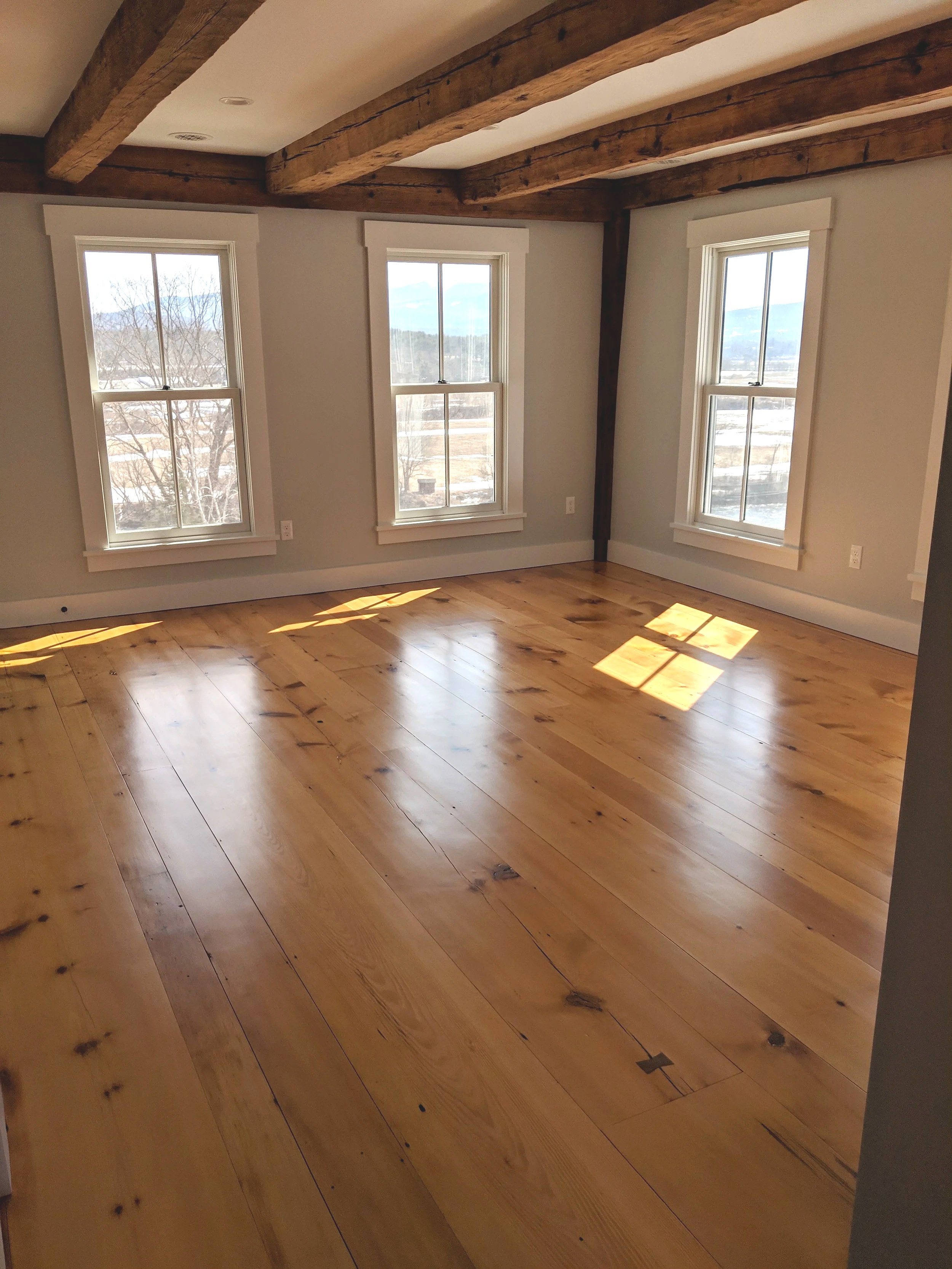 Empty room with three large windows, wooden floor, white walls, and exposed wooden beams on the ceiling.