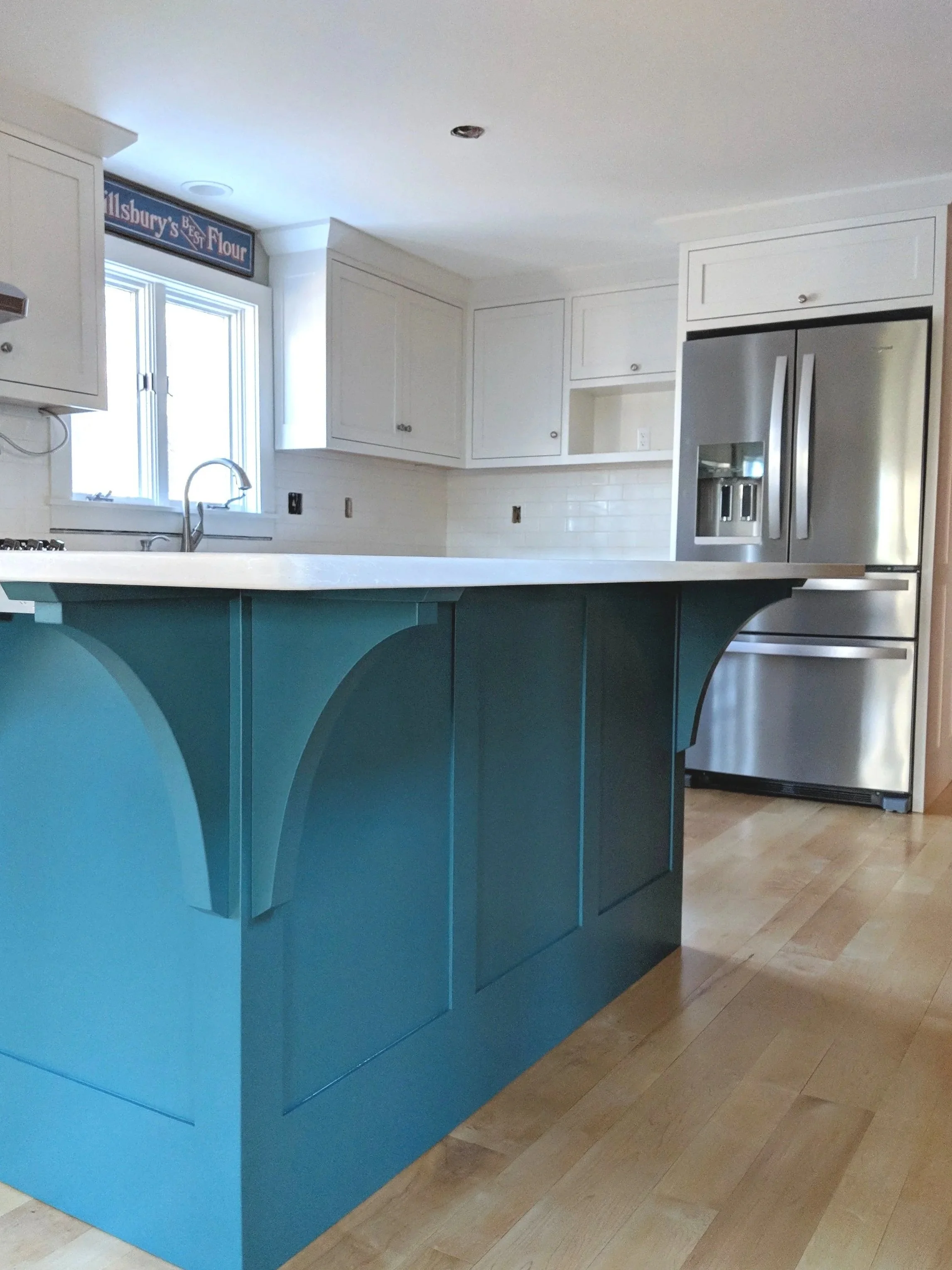Modern kitchen with white cabinets, stainless steel refrigerator, teal island with decorative paneling, and light wooden floor.