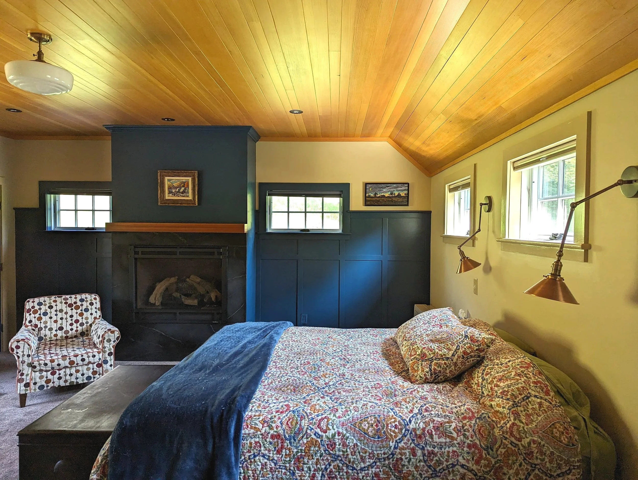 A cozy bedroom with a wooden vaulted ceiling, a bed with a floral bedspread, a blue blanket, and a matching pillow. There is a patterned armchair near a fireplace with black paneling and two small windows, along with three small windows on the right 