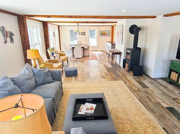 View of a spacious living room with a sofa, armchair, coffee table, and a fireplace, leading into a dining area with a round table and chairs, featuring wooden flooring and large windows letting in natural light.