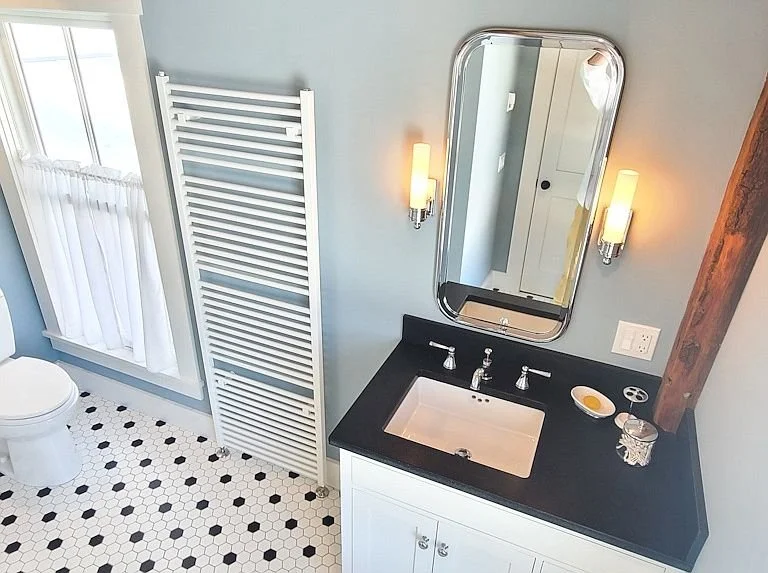 Bathroom with black countertop vanity, oval mirror, two wall sconces, white sink, toilet near window with white curtains, white towel radiator, and patterned black and white floor tiles.