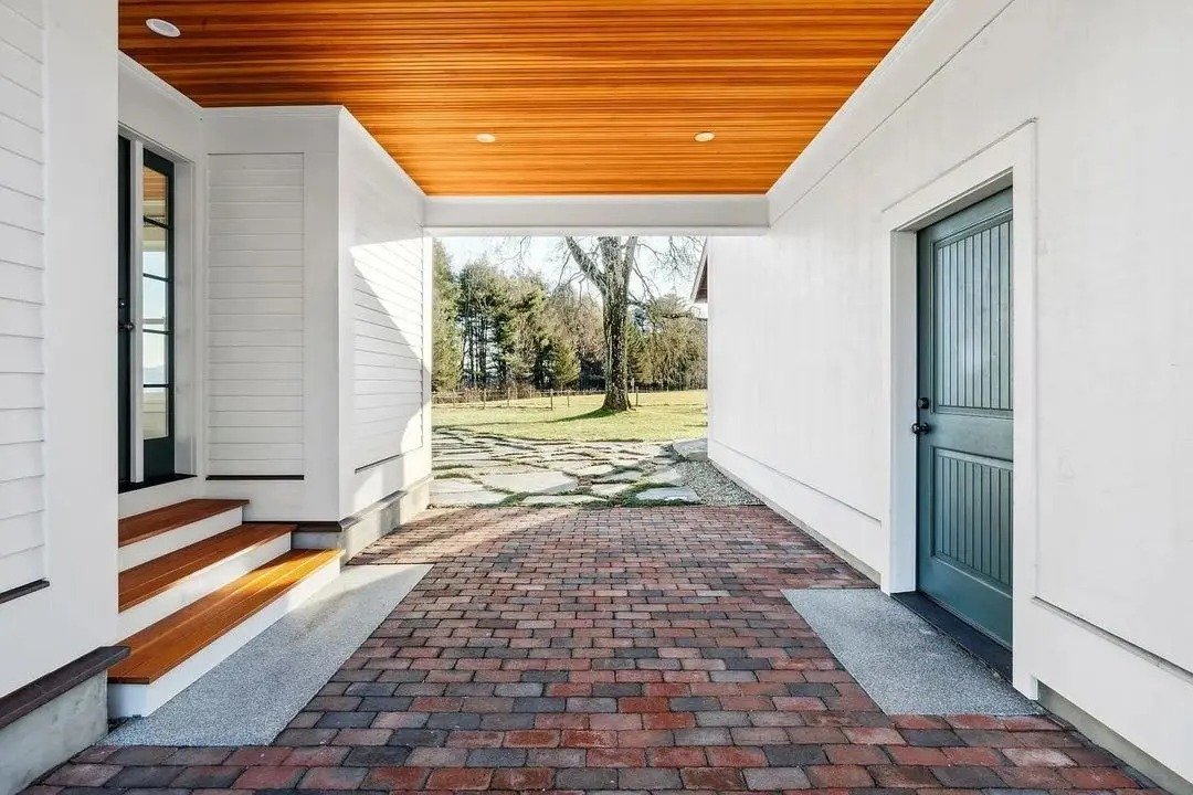 Covered porch area with brick flooring, white siding walls, a wooden ceiling with recessed lighting, a set of stairs, a blue door, and an open view of an outdoor yard with trees.