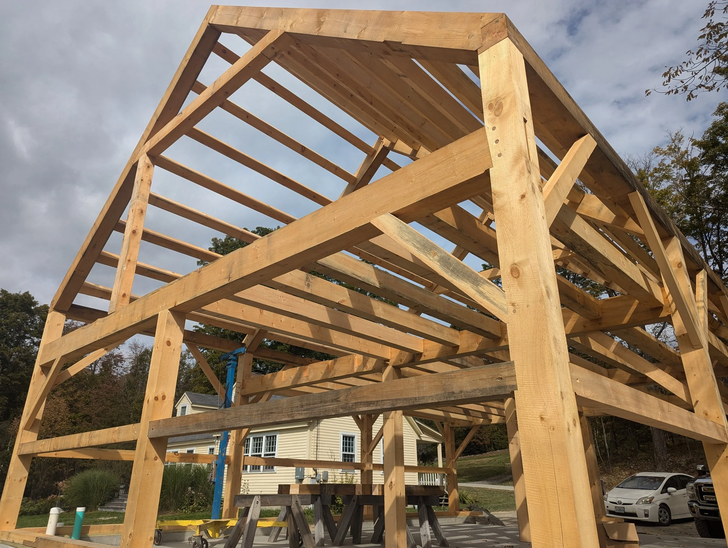 A wooden house frame under construction with multiple floors, set outdoors near a house, with trees in the background and a car parked on the street.