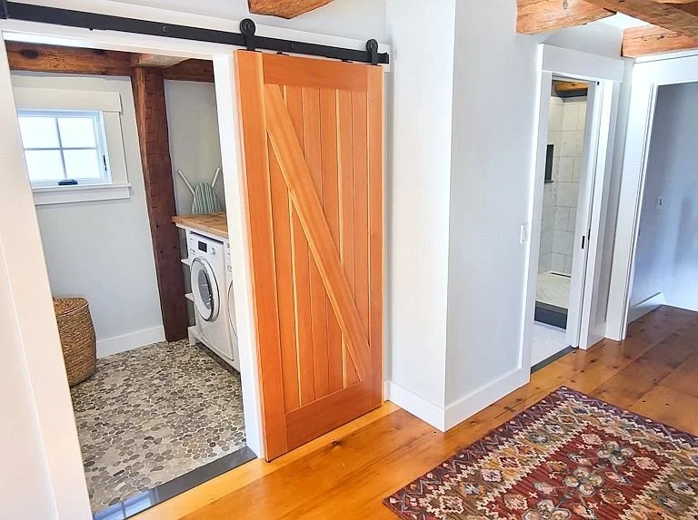 Interior view of a home showing a wood sliding barn door, laundry nook with a washing machine, and a bathroom with a shower. Hardwood floors and a patterned area rug are visible.