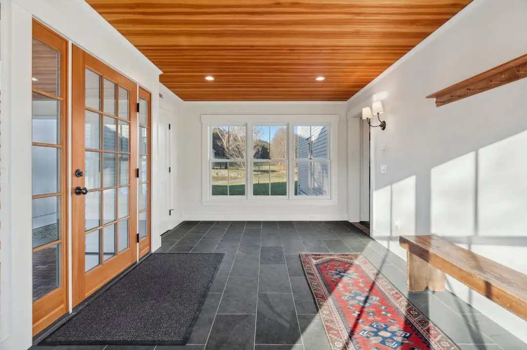 Living room with large window, wood ceiling, French doors, black tile floor, white walls, wall-mounted lamps, wooden bench, patterned rug, and outdoor greenery view.