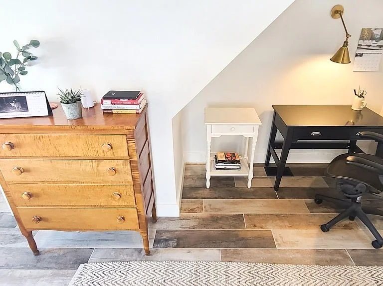 Interior of a home office with a wooden dresser, a small white side table, a black desk, a swivel chair, and a brass wall-mounted lamp.