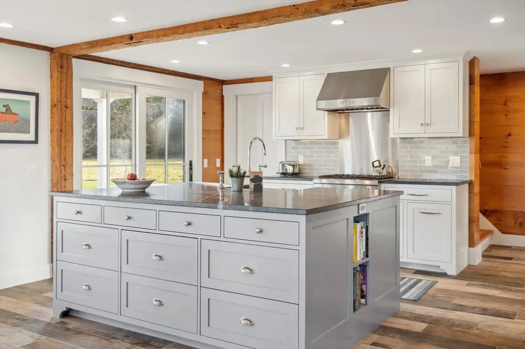 Modern kitchen with white cabinets, gray island, stainless steel range hood, wooden accents, large window, and hardwood floors.