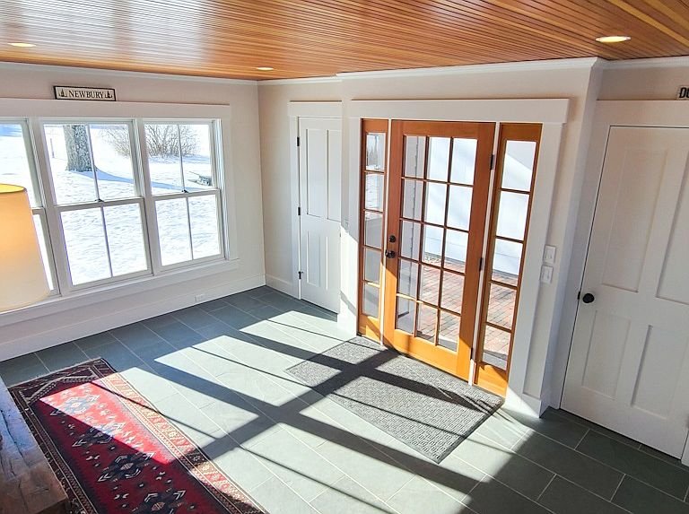 Bright room with large window, wooden French doors, and tiled floor casting shadows, with white and wooden interior.