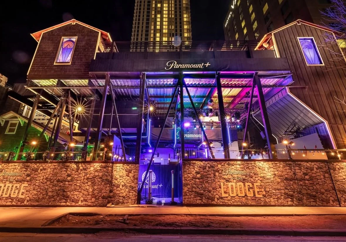 Night view of a multi-level building with neon lights, labeled 'Paramount+' and 'The Lodge,' featuring a stone wall and outdoor seating area.