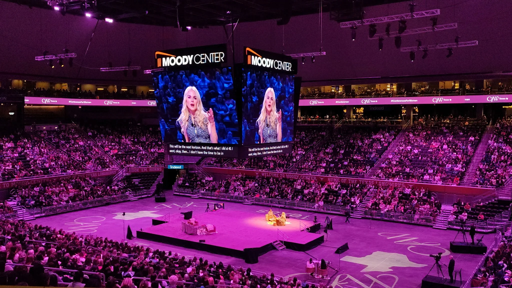Large indoor arena filled with audience watching a discussion on stage, with two large screens displaying a woman speaking and subtitles, and purple lighting throughout.