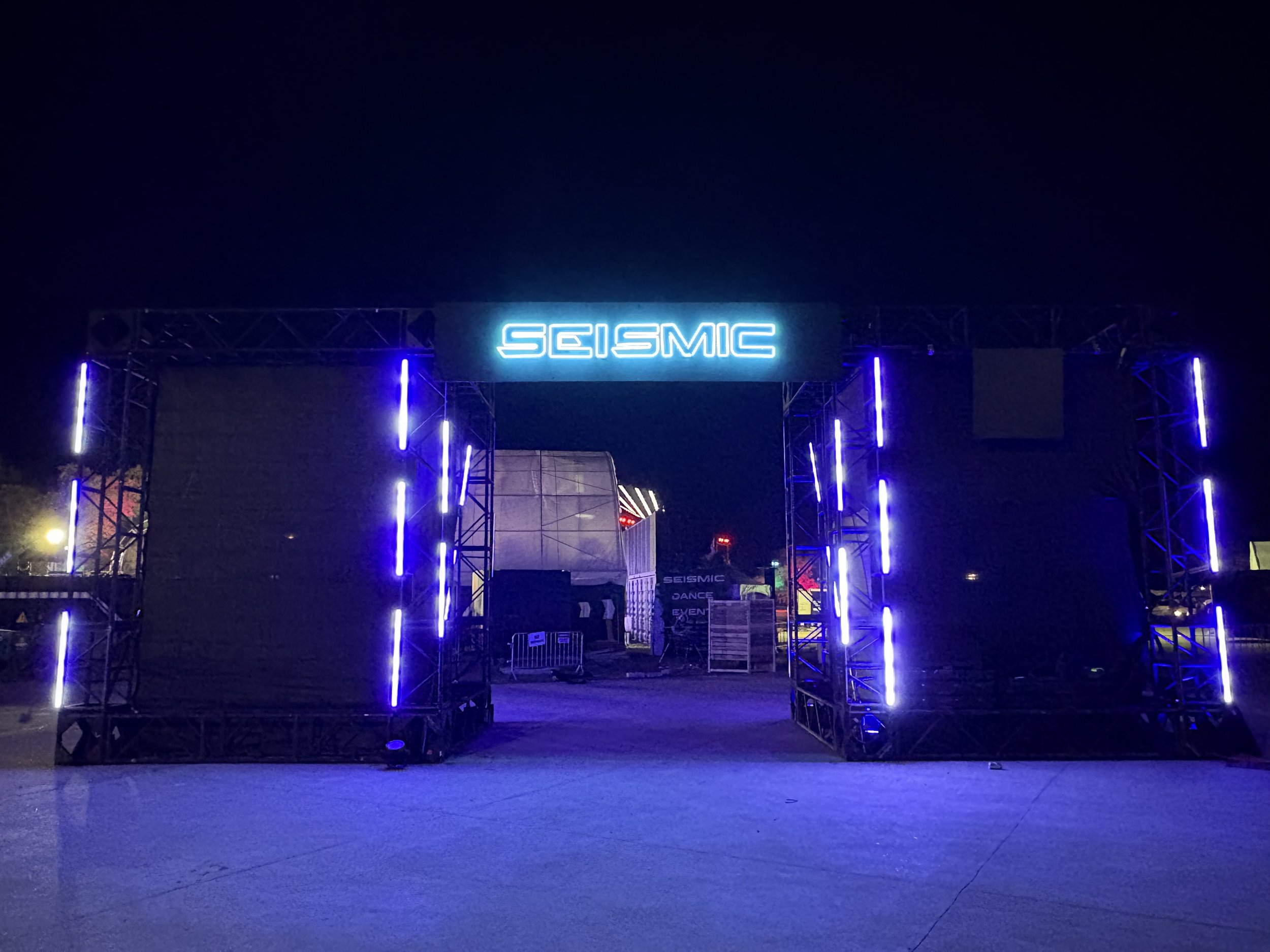 Nighttime view of a large illuminated sign that reads 'SEISMIC' with blue neon lights, flanked by black speakers or sound equipment.