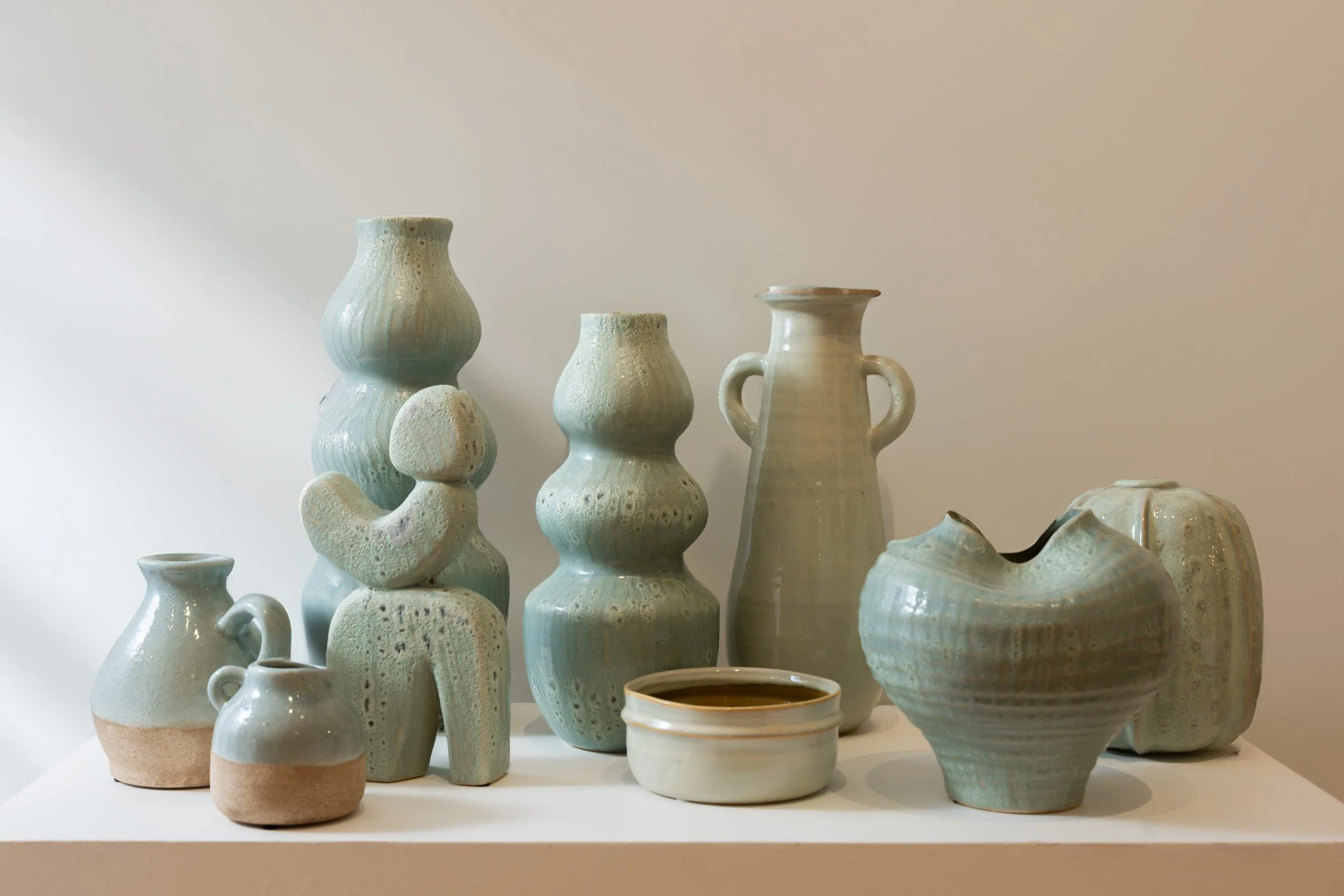 Collection of ceramic vases and pots in various shapes and sizes, with a light turquoise glaze, arranged on a white surface against a plain light wall.