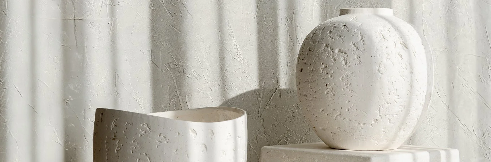 Two white textured ceramic vases, one with a rounded shape and the other with a rectangular opening, are placed on a white surface against a light-colored textured wall with shadows.