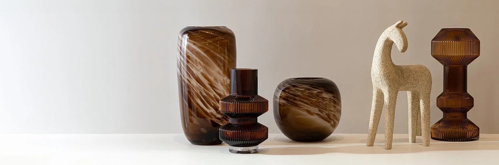 Collection of decorative vases and a sculpture on a white surface against a neutral wall. Includes tall and short brown marble-patterned glass vases, a textured ceramic horse sculpture, and geometrically shaped glass vases.