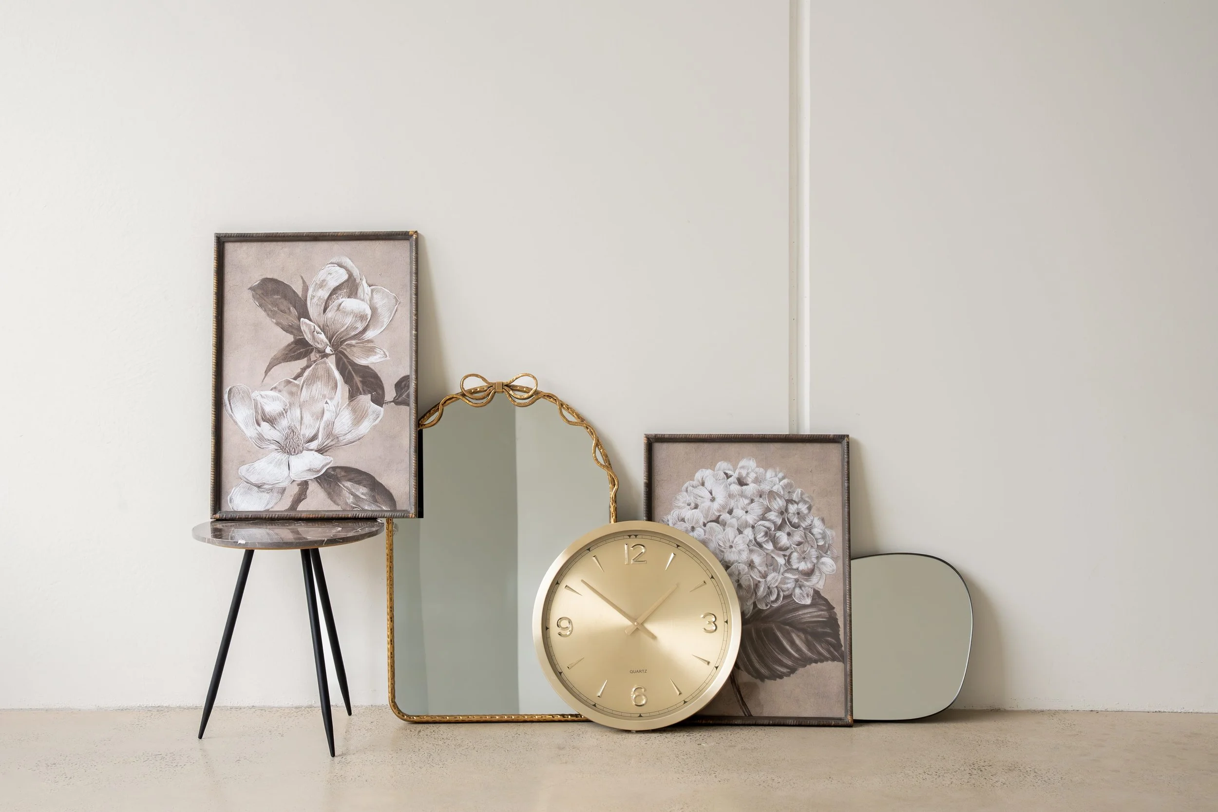 Decorative arrangement with framed botanical prints, a gold-framed mirror, a gold clock, and a small table with black legs against a white wall.