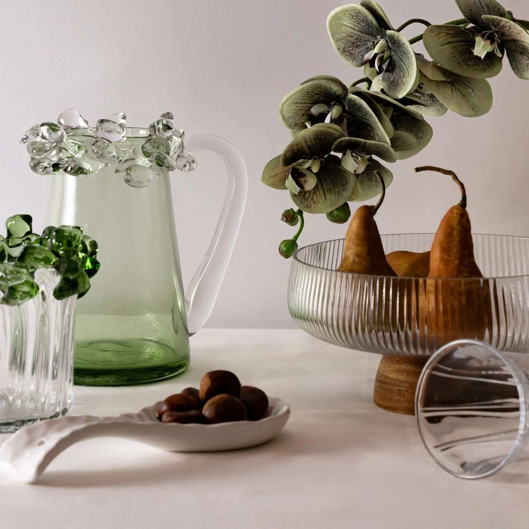 Close-up of a clear glass pitcher with a handle, filled with water and decorative glass stones on top, and a white ceramic bowl with some brownish fruit, possibly figs or dates, beside it. In the background, there is a plant with green and white variegated leaves in a metal wire basket.