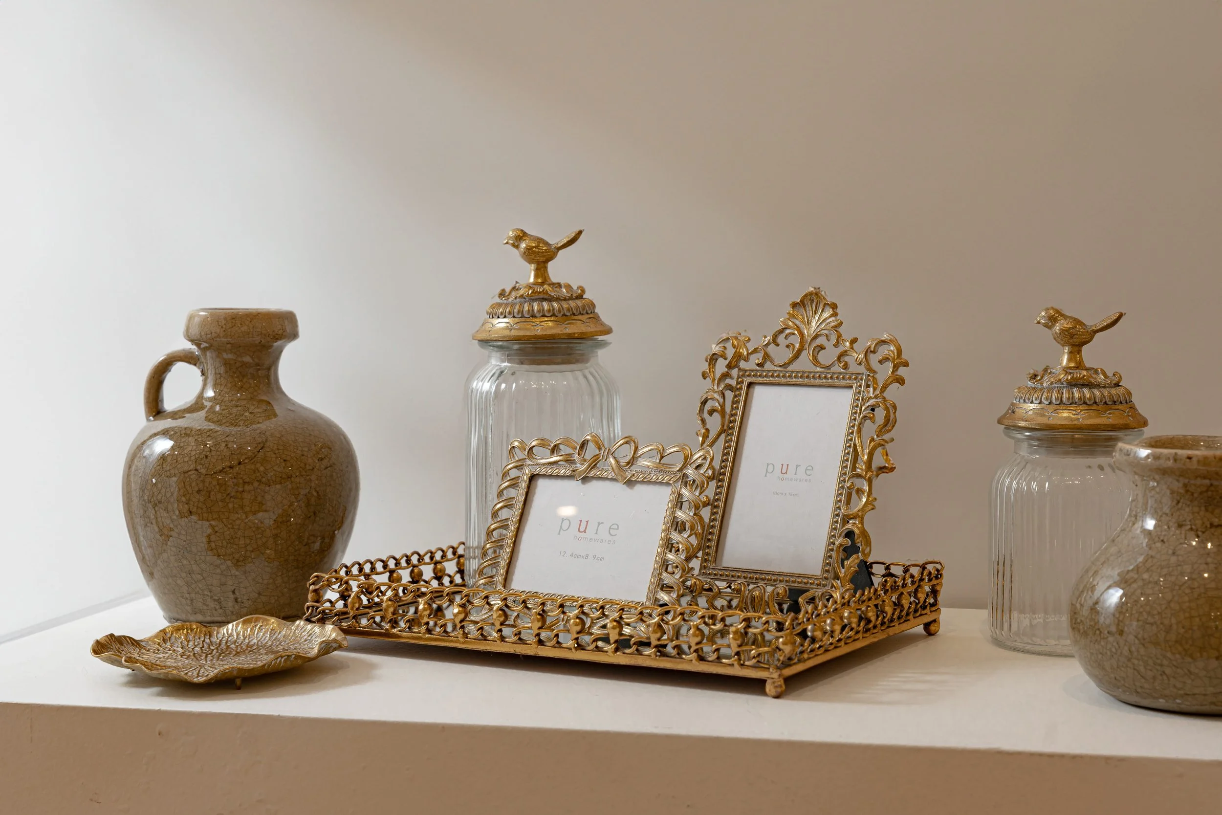 A display of decorative items including two glass containers with gold bird toppers, ornate gold picture frames, a ceramic vase, a gold-colored tray, and a decorative gold tray in a white setting.
