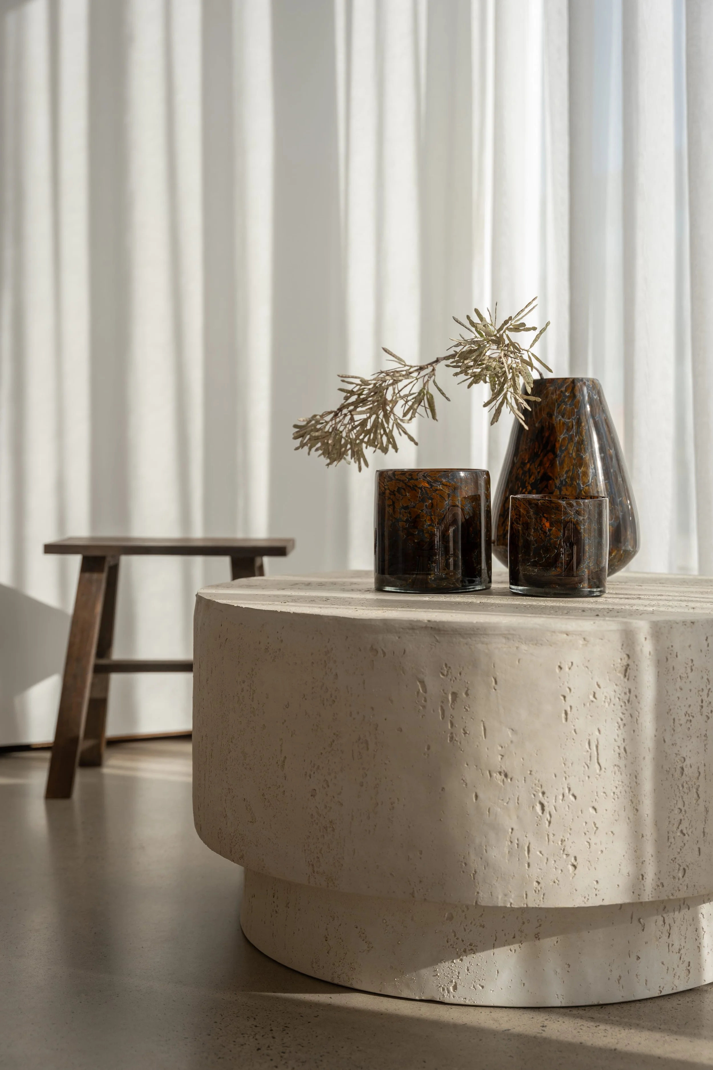 Decorative items on a round table, including a large brown vase with a branch, and two smaller matching vases, with a background of white curtains.