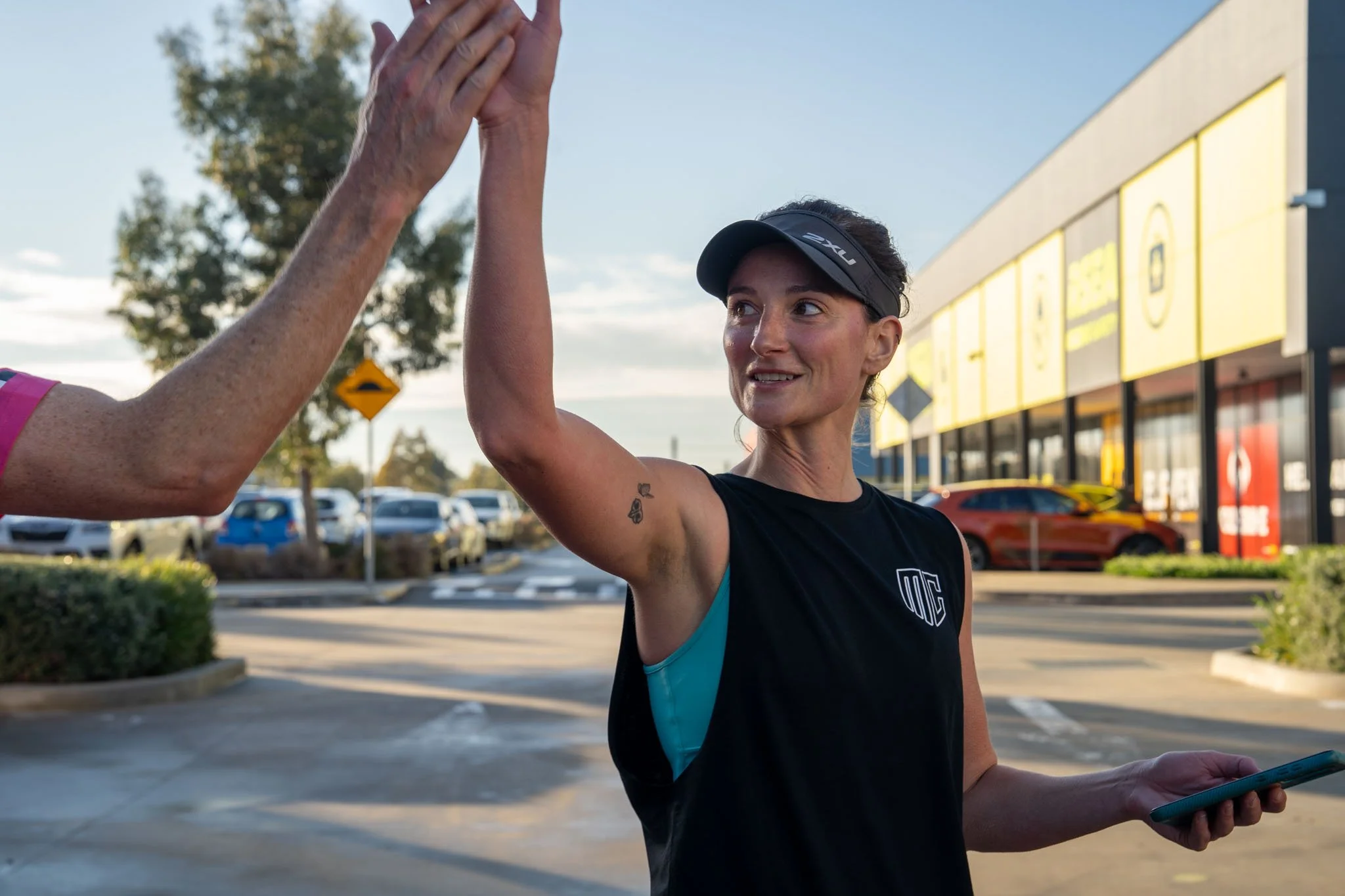 A woman wearing a black sports top and a black visor is giving a high-five to another person in a parking lot outside a retail store during the daytime. She is holding a smartphone in her left hand and has a tattoo of three stars on her right arm.
