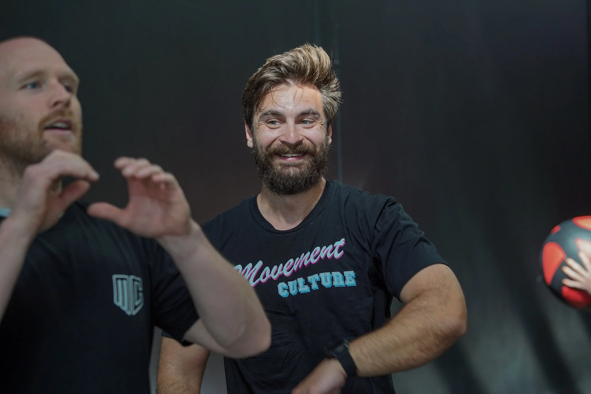 A man sitting on a padded box with the words 'NE FITNESS' on it, watching a man in black athletic clothing performing a stretch or exercise. The background features a black wall with the words 'MOTION CULTURE' in stylized blue and pink outline text.