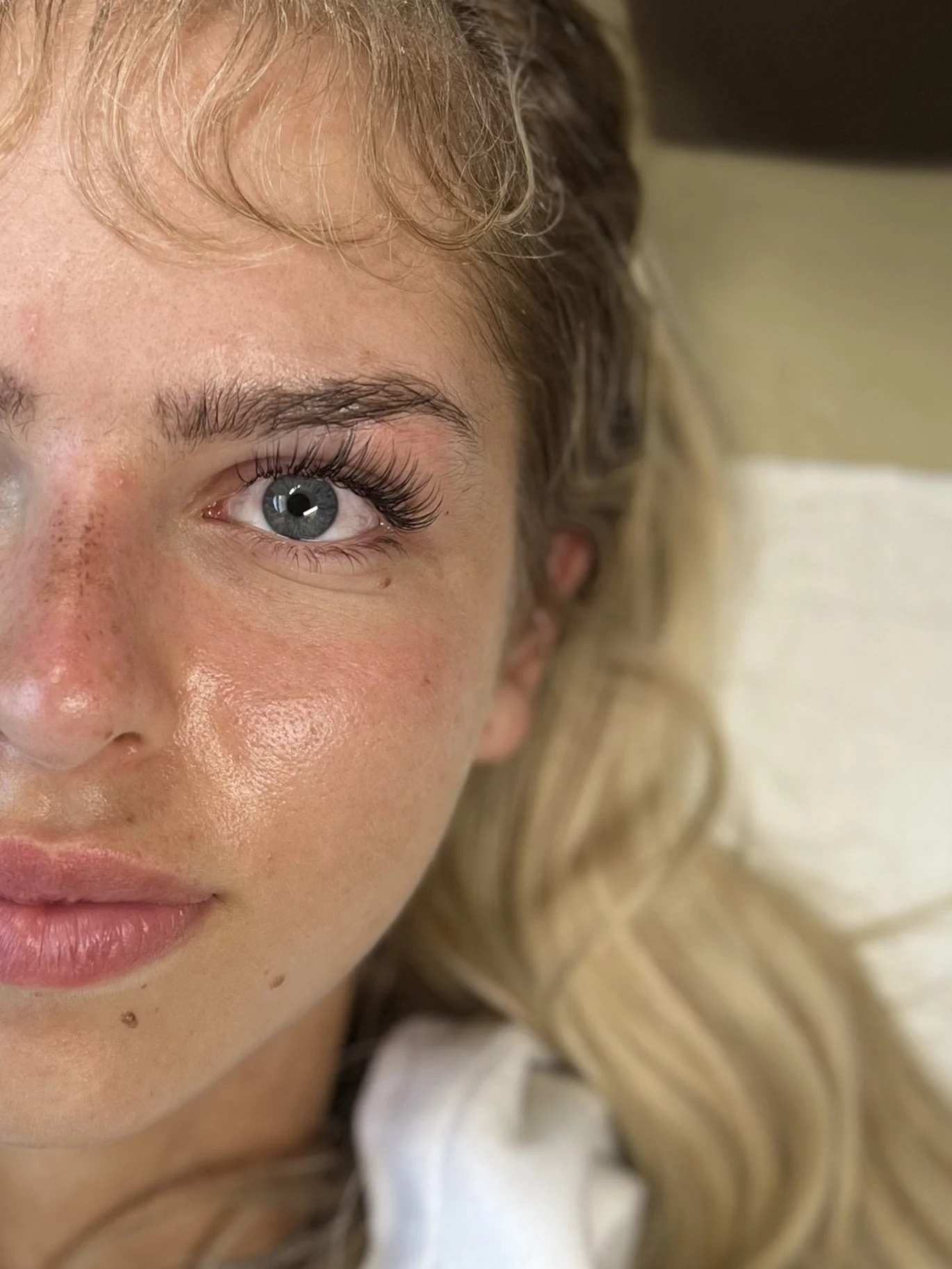 Close-up of a young woman's face, showing her eye, cheek, and part of her lips, with blonde wavy hair and natural makeup.