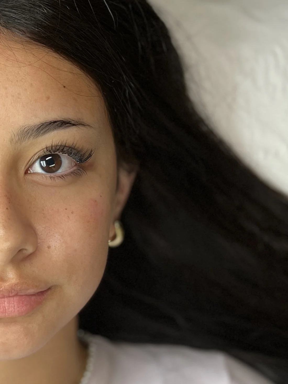 Close-up of a young woman's face, showing her left eye, part of her nose, cheek, and lips, with dark hair and a gold hoop earring.