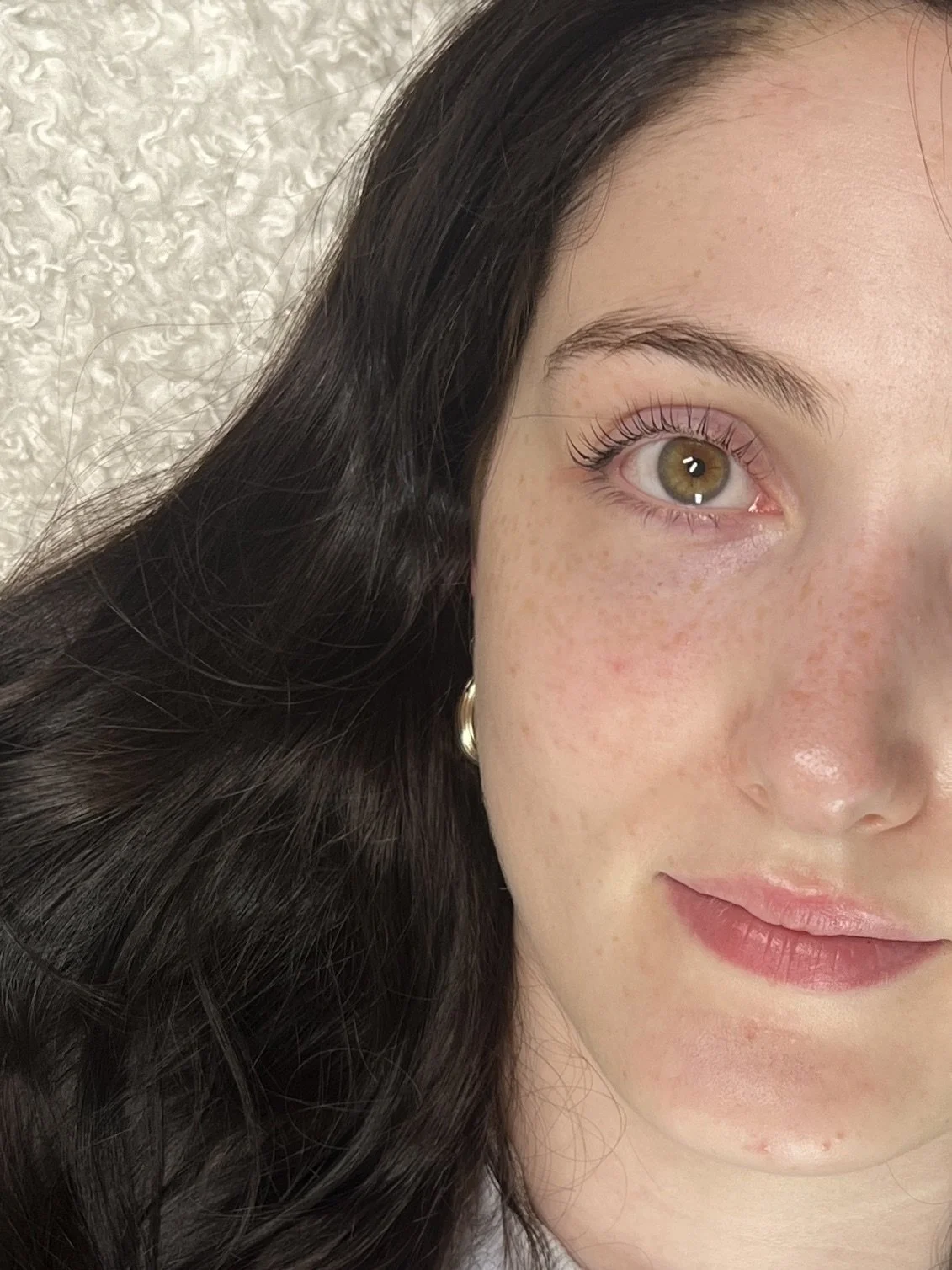 Close-up of a young woman's face showing her eye, cheek, and lips, with dark brown hair and a small hoop earring.