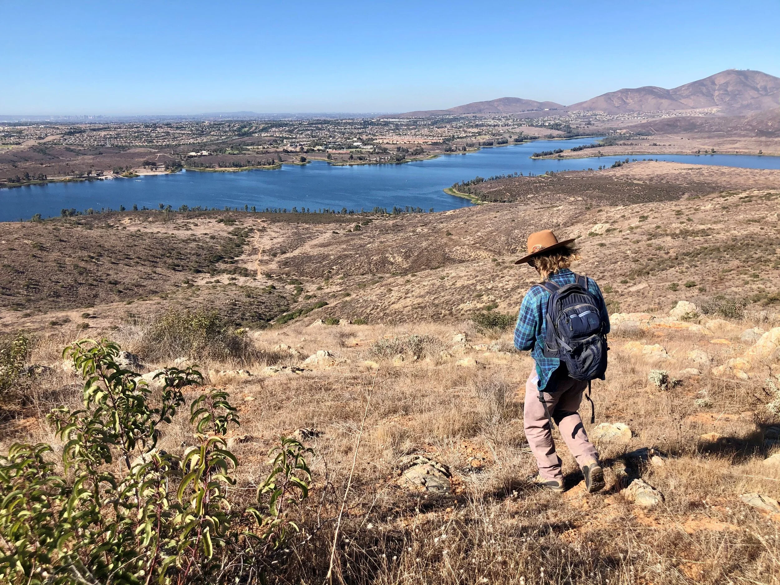 A person wearing a hat and backpack walking on a dry hill, overlooking a blue lake with distant mountains and a clear sky.